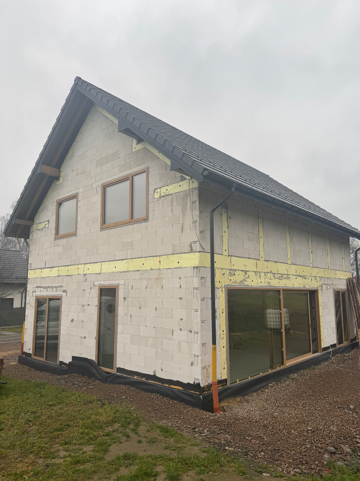 Surowy stan domu jednorodzinnego z betonu komórkowego z zamontowanymi oknami i czarną membraną hydroizolacyjną wokół fundamentów, widoczne żółte elementy ocieplenia między blokami, dach pokryty...