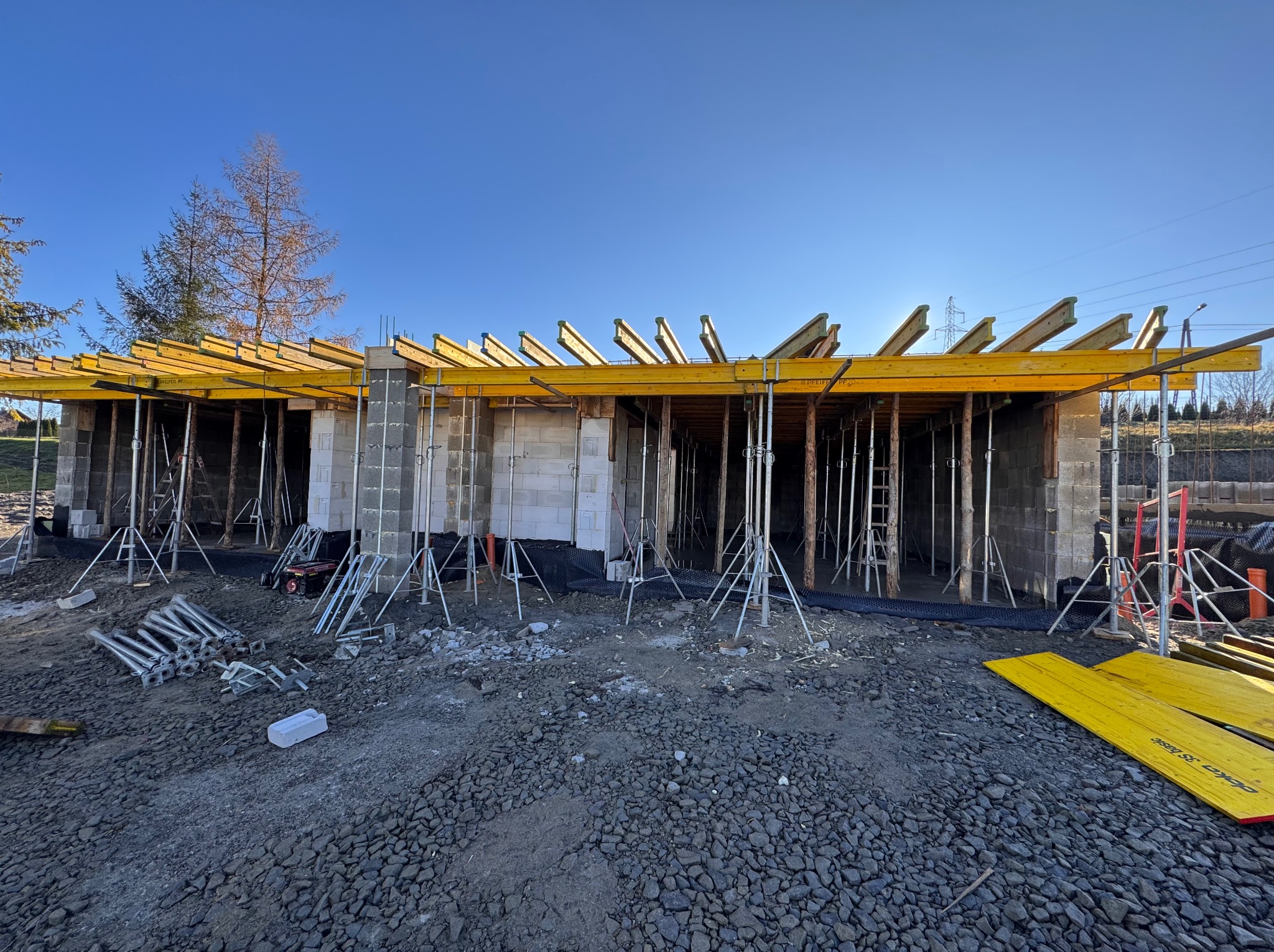 Budowa domu z bloczków betonowych, szalunki stropowe z żółtymi belkami, podpory stropowe, widok na plac budowy w słoneczny dzień.