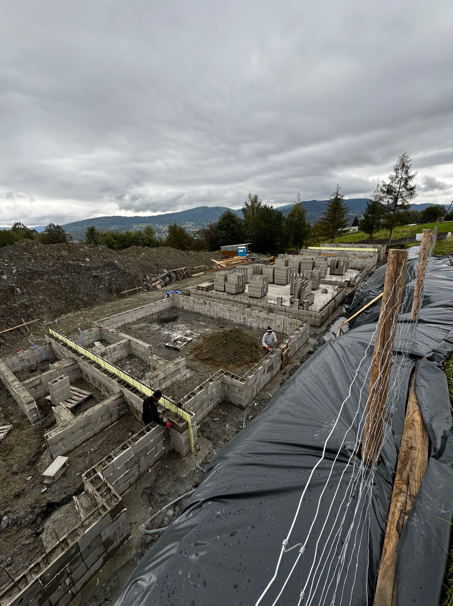 Wykopane fundamenty domu z bloczków betonowych, zbrojenie stalowe wystaje, widoczna izolacja pionowa i pracownicy na budowie, w tle górzysty krajobraz pod pochmurnym niebem.