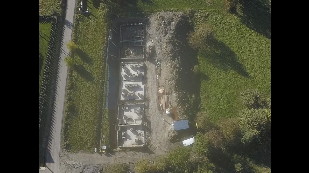 Widok z góry na plac budowy z przygotowanymi fundamentami pod budynek, widoczne szalunki i zbrojenia, obok składowisko materiałów budowlanych i zaparkowany kontener biurowy.