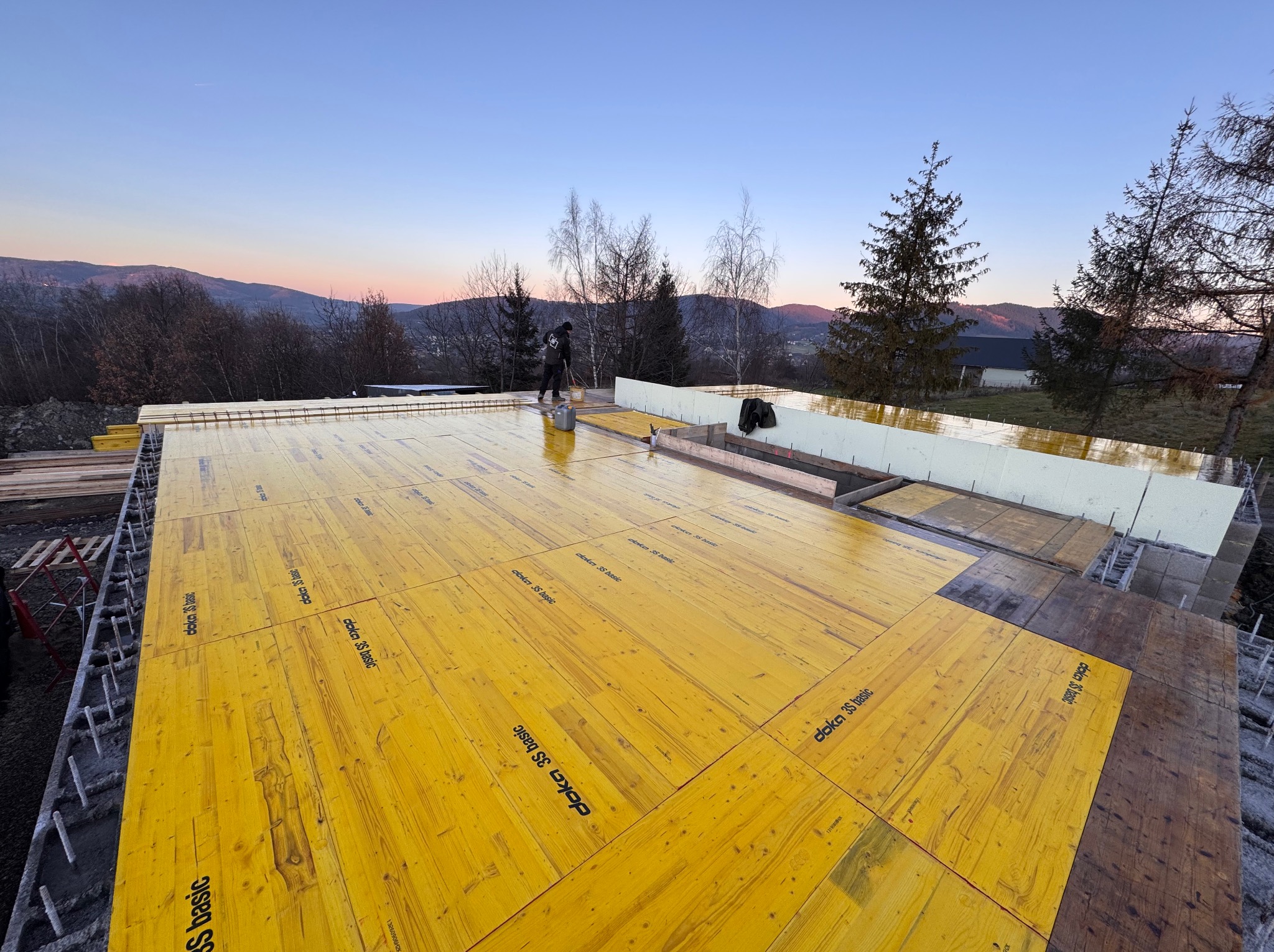 Wylewanie fundamentów pod budowę domu jednorodzinnego na tle górskiego krajobrazu o zmierzchu. Żółte płyty szalunkowe z logo Doka 3S Basic ułożone na zbrojeniu.