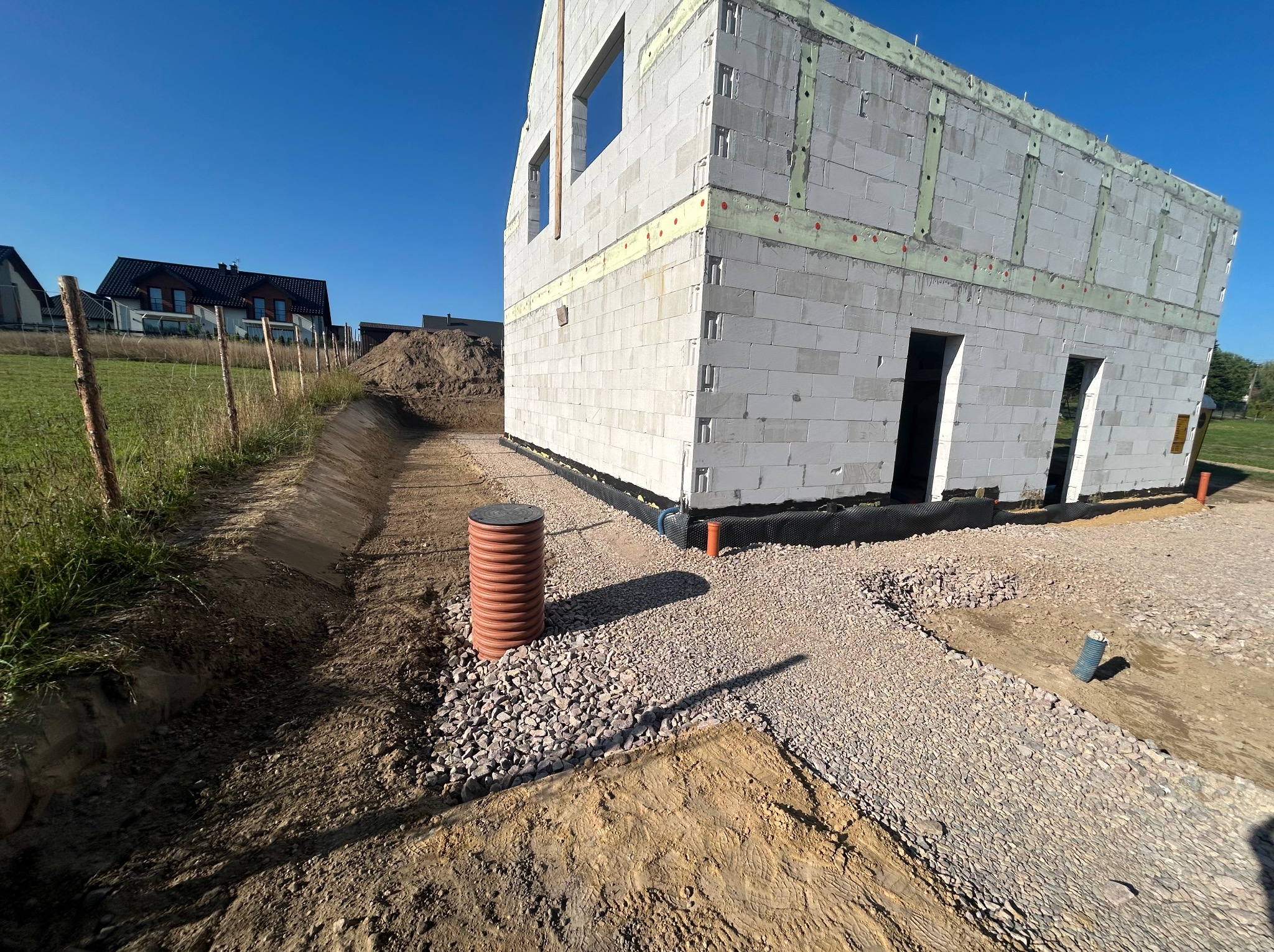 Surowy stan domu z białych bloczków, widoczne otwory okienne i drzwiowe, teren przygotowany, częściowo utwardzony żwirem, na pierwszym planie studzienka kanalizacyjna z tworzywa sztucznego.