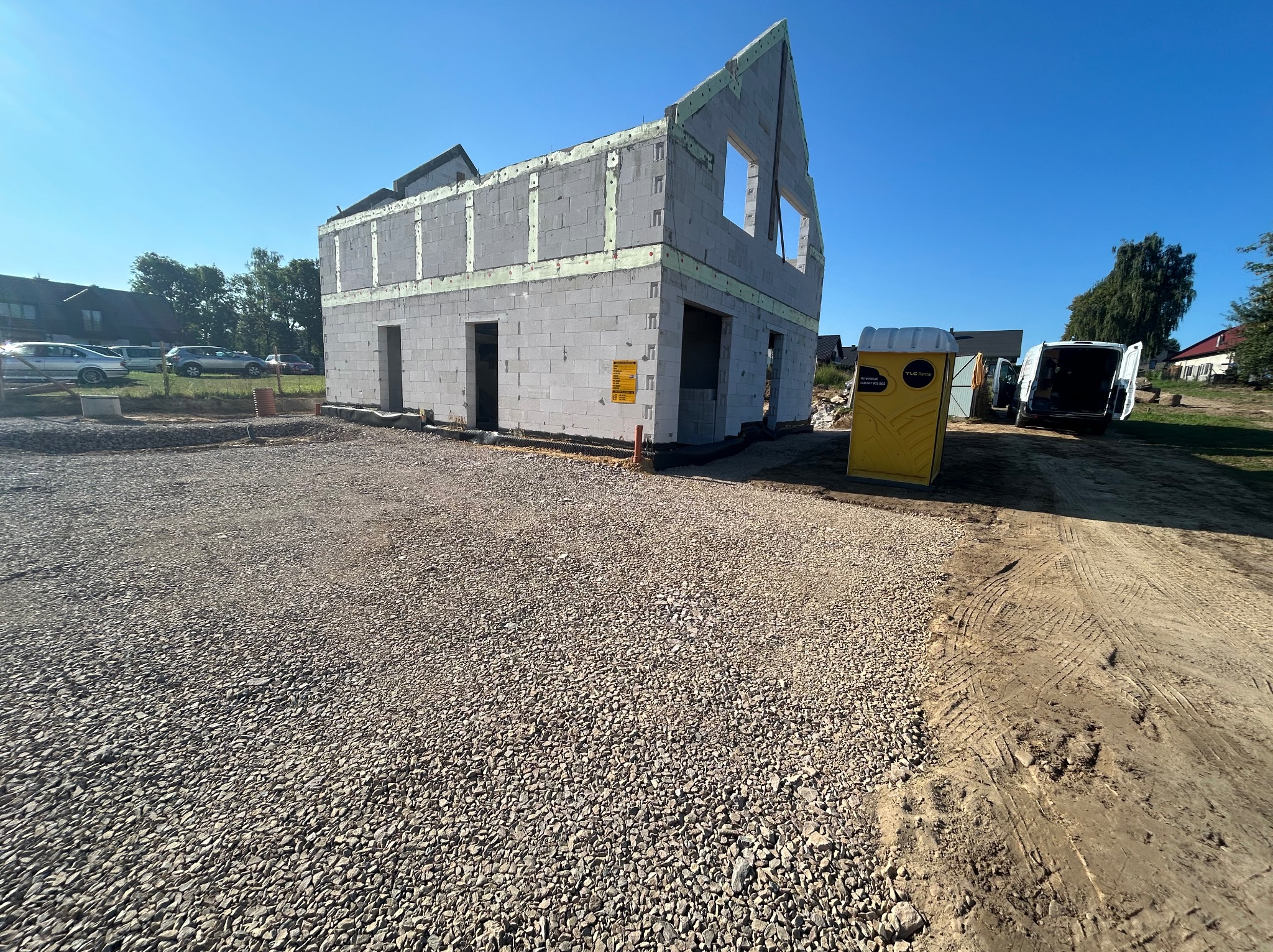 Surowy stan dwukondygnacyjnego domu murowanego z betonu komórkowego w trakcie budowy, z widocznymi otworami okiennymi i drzwiowymi, ociepleniem na obwodzie i żółtą toaletą przenośną na placu budowy.