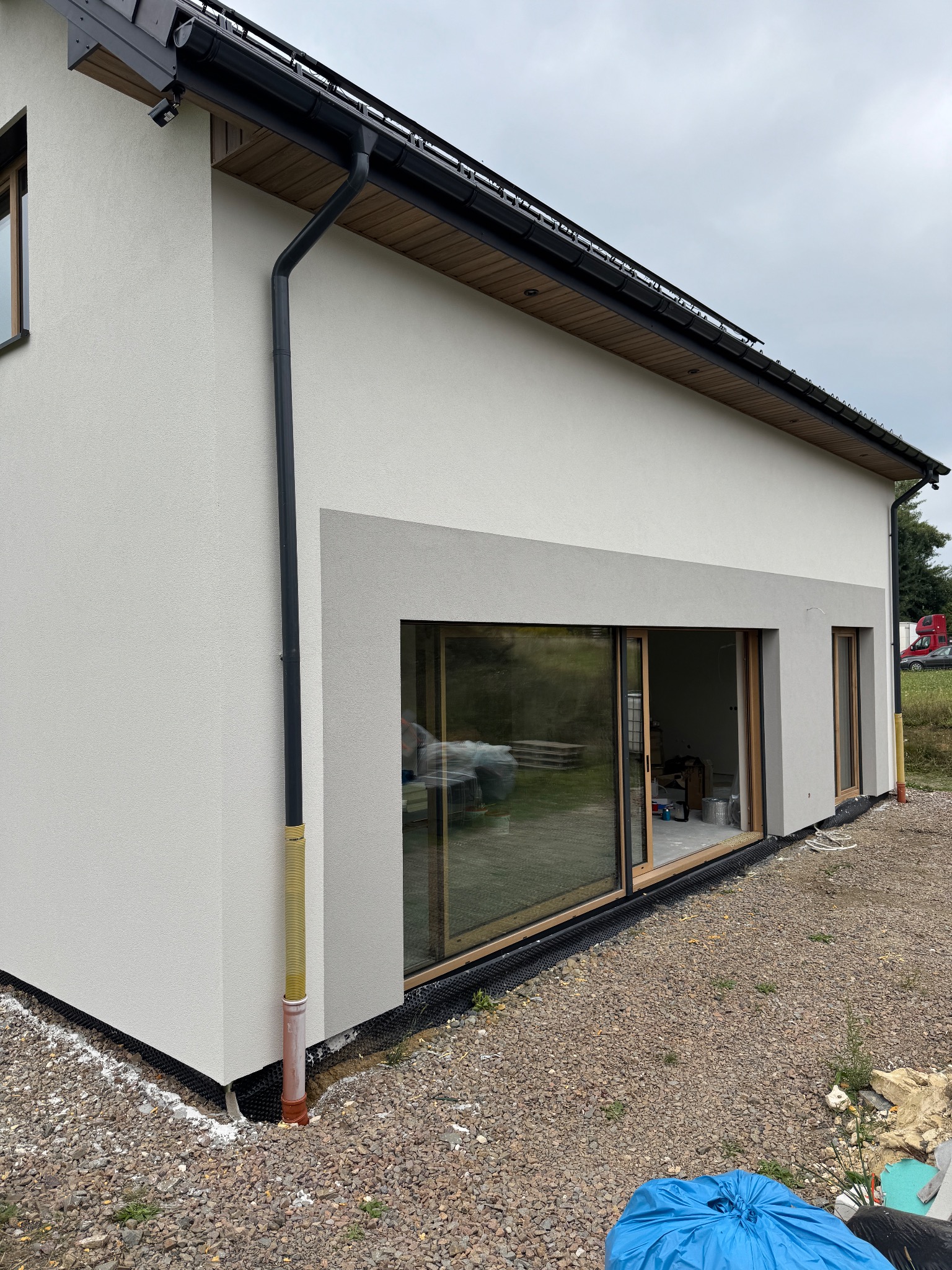 Nowoczesny dom jednorodzinny z dużym oknem przesuwnym i szarą elewacją, widok z zewnątrz. Widoczne detale wykończenia i otoczenie budynku.