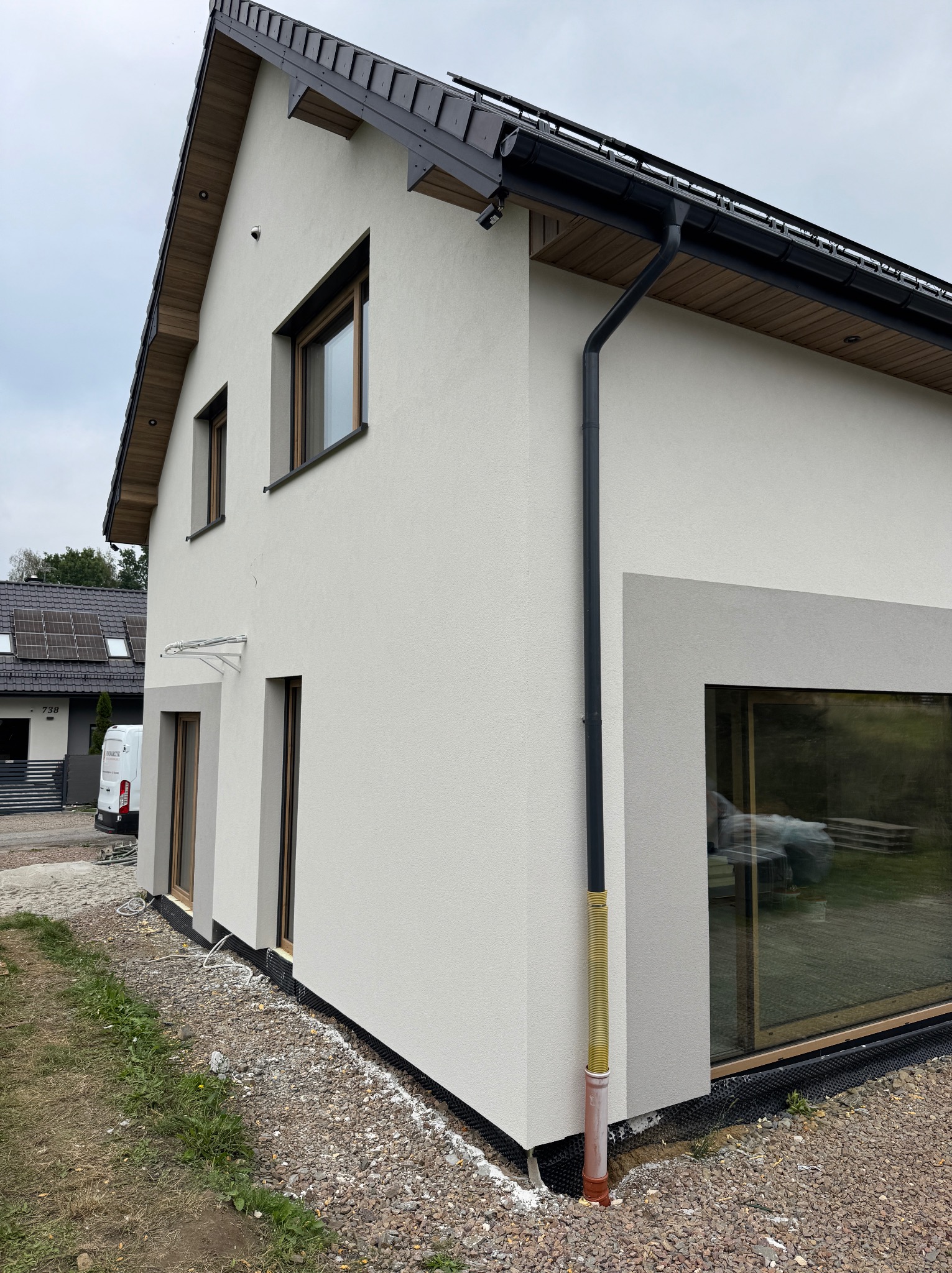 Nowoczesny dom z jasną elewacją, ciemnym dachem i dużym oknem narożnym. Widoczny system rynnowy i fragment ogrodu. Minimalistyczny design z elementami wykończenia drewnem.