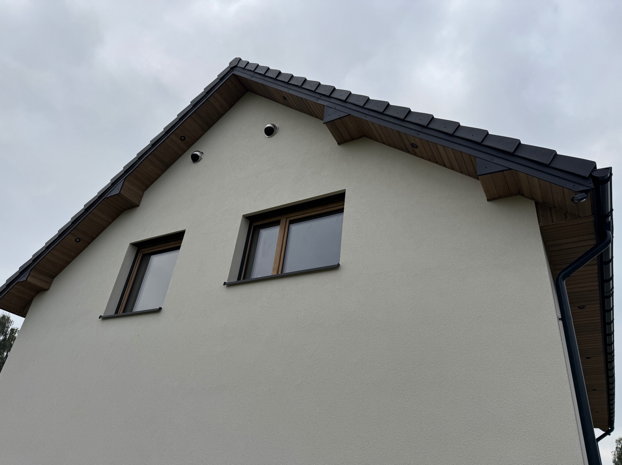 Fragment elewacji domu z dwoma oknami, czarną dachówką i rynną. Jasna elewacja o fakturze baranka. Widok z dołu na szczyt budynku pod pochmurnym niebem.