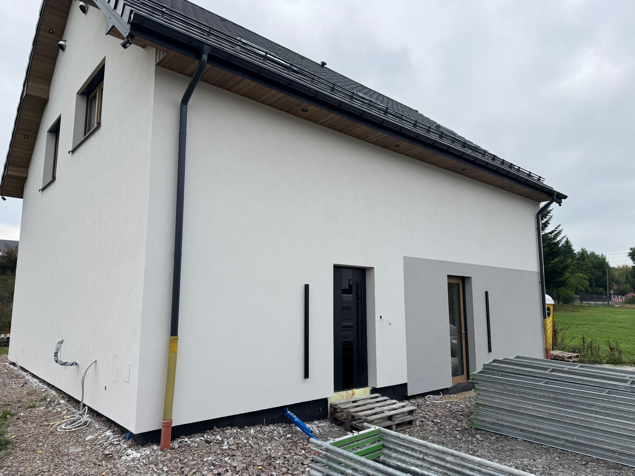 Nowoczesny dom jednorodzinny z jasną elewacją i ciemną stolarką okienną. Widoczne detale wykończenia, rynny i fragment dachu. Na pierwszym planie materiały budowlane.