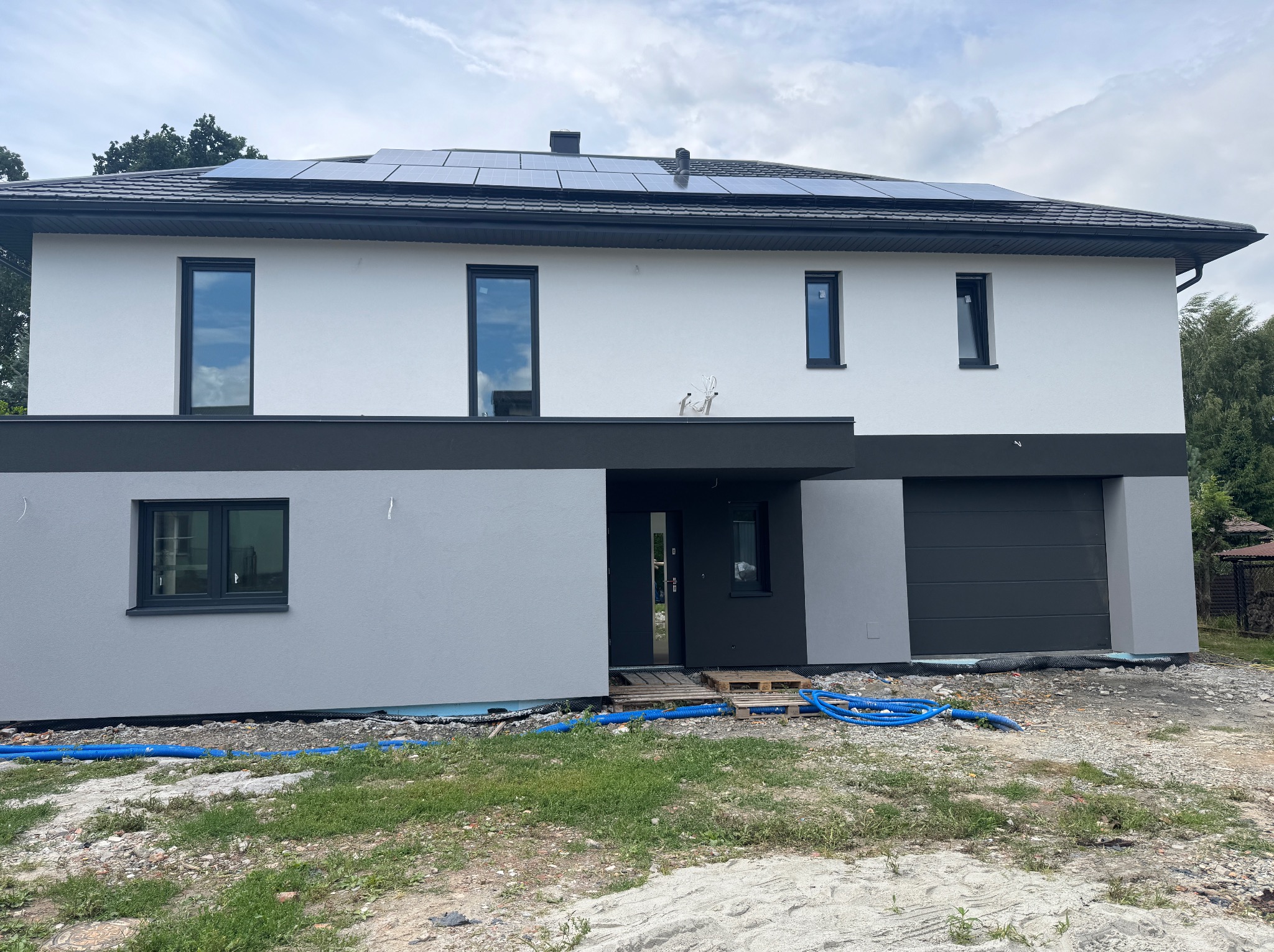 Dwukondygnacyjny dom z panelami słonecznymi na dachu, w trakcie wykończenia elewacji. Widoczne niebieskie rury na trawniku i paleta pod drzwiami.
