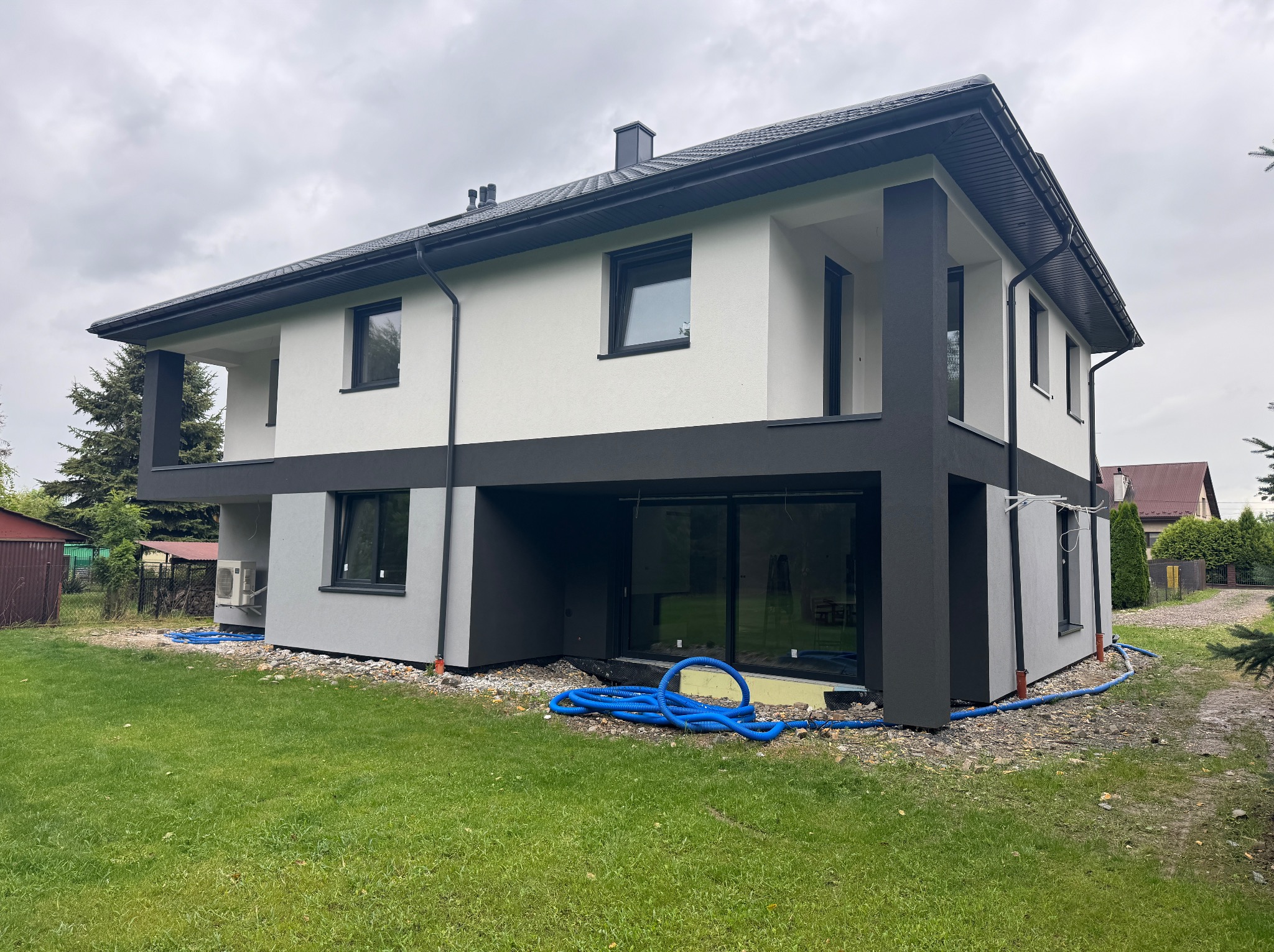 Dwukondygnacyjny dom z płaskim dachem w odcieniach szarości i bieli, z widoczną instalacją wokół budynku na trawniku. Nowoczesna architektura, pochmurne niebo.