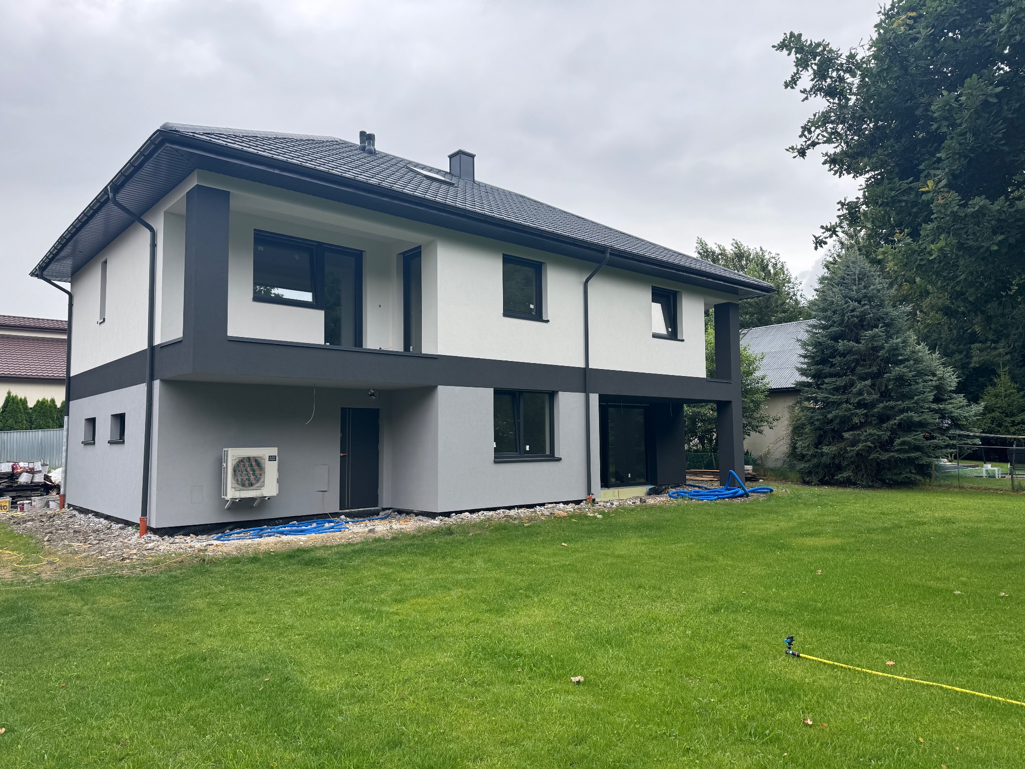 Dwukondygnacyjny dom z płaskim dachem, pomalowany na biało i szaro, z widocznym klimatyzatorem na elewacji i zielonym trawnikiem. Ujęcie z boku, w tle drzewa.