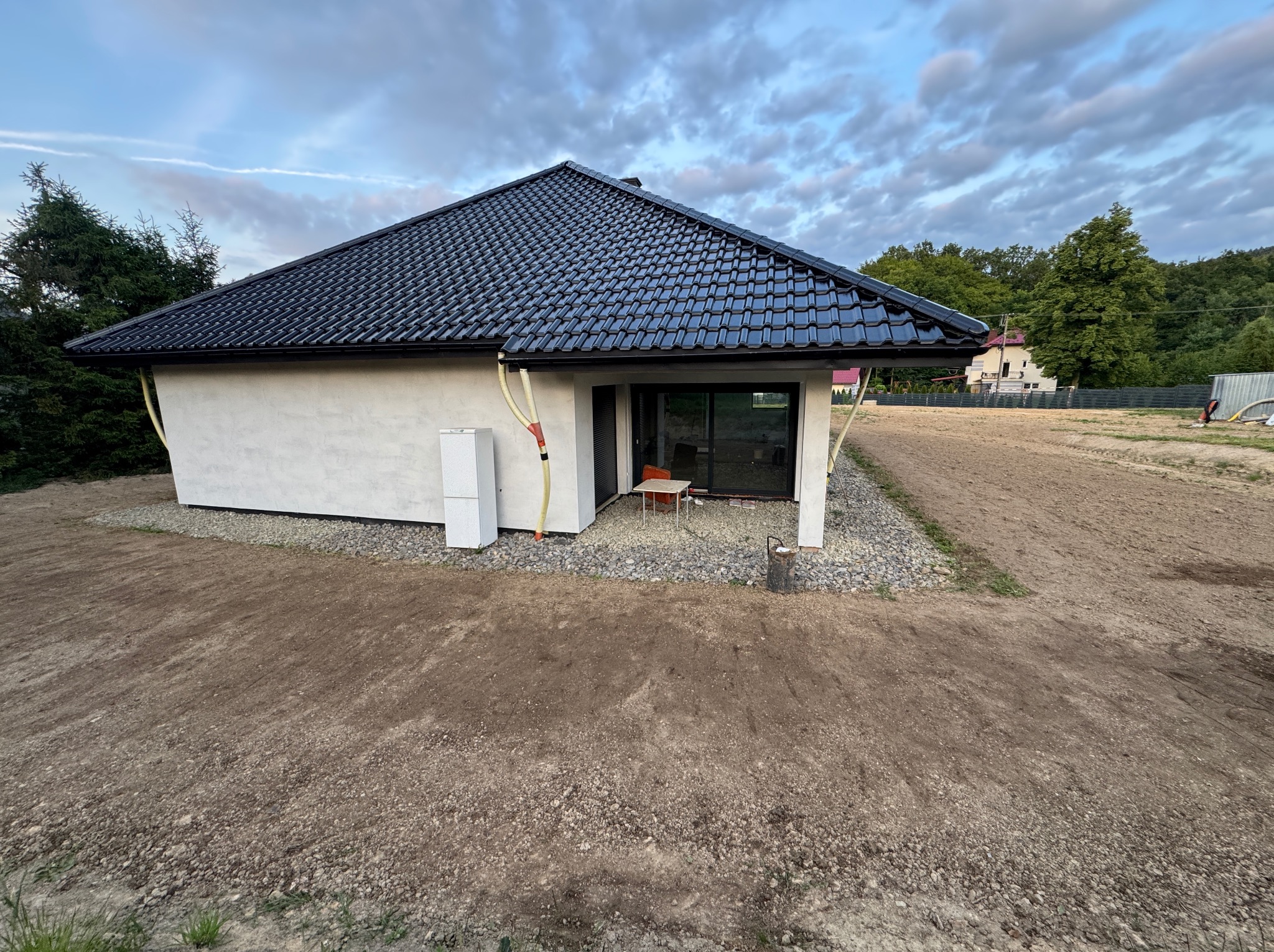 Nowoczesny, parterowy dom z czarną dachówką i białymi ścianami, otoczony ziemią i kamieniami. Widoczne okno tarasowe i instalacje na elewacji. Ujęcie z zewnątrz.