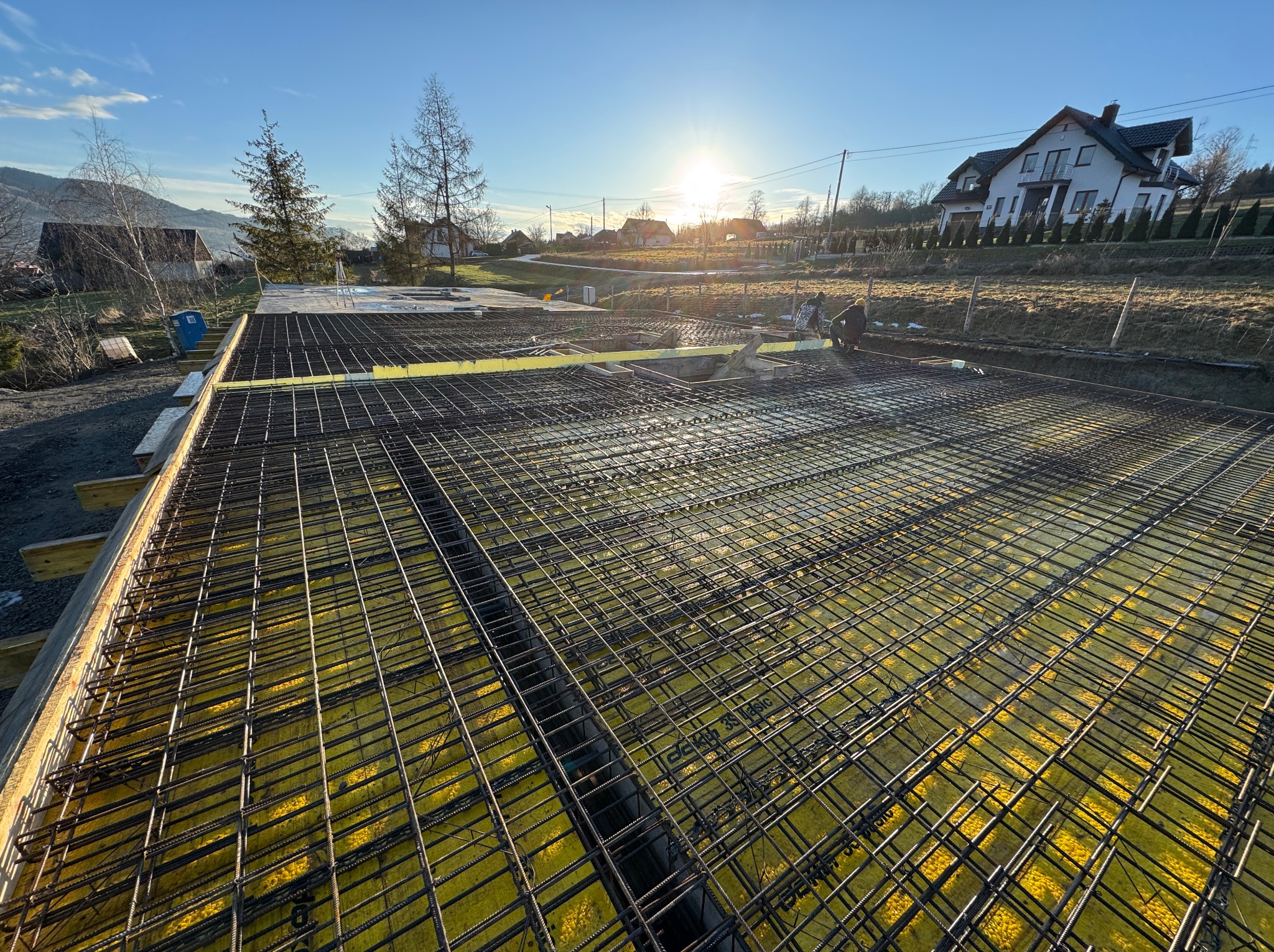 Zbrojenie fundamentu domu jednorodzinnego z żółtą izolacją, widoczne elementy szalunku i pracownicy w tle, w słoneczny dzień na tle górskiego krajobrazu.