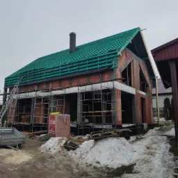 Surowy stan budowy domu jednorodzinnego z częściowo wykonaną konstrukcją dachu, widoczne rusztowania i materiały budowlane na zewnątrz w zimowej scenerii.