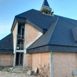 Niedokończona budowa kościoła z nowym, czarnym pokryciem dachowym z blachy trapezowej, widoczne rusztowania i surowy stan ścian z czerwonej cegły.