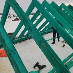 Zielona, drewniana konstrukcja szkieletu dachu w trakcie montażu, widoczny pracownik w roboczych spodniach i narzędzia na śnieżnym podłożu.