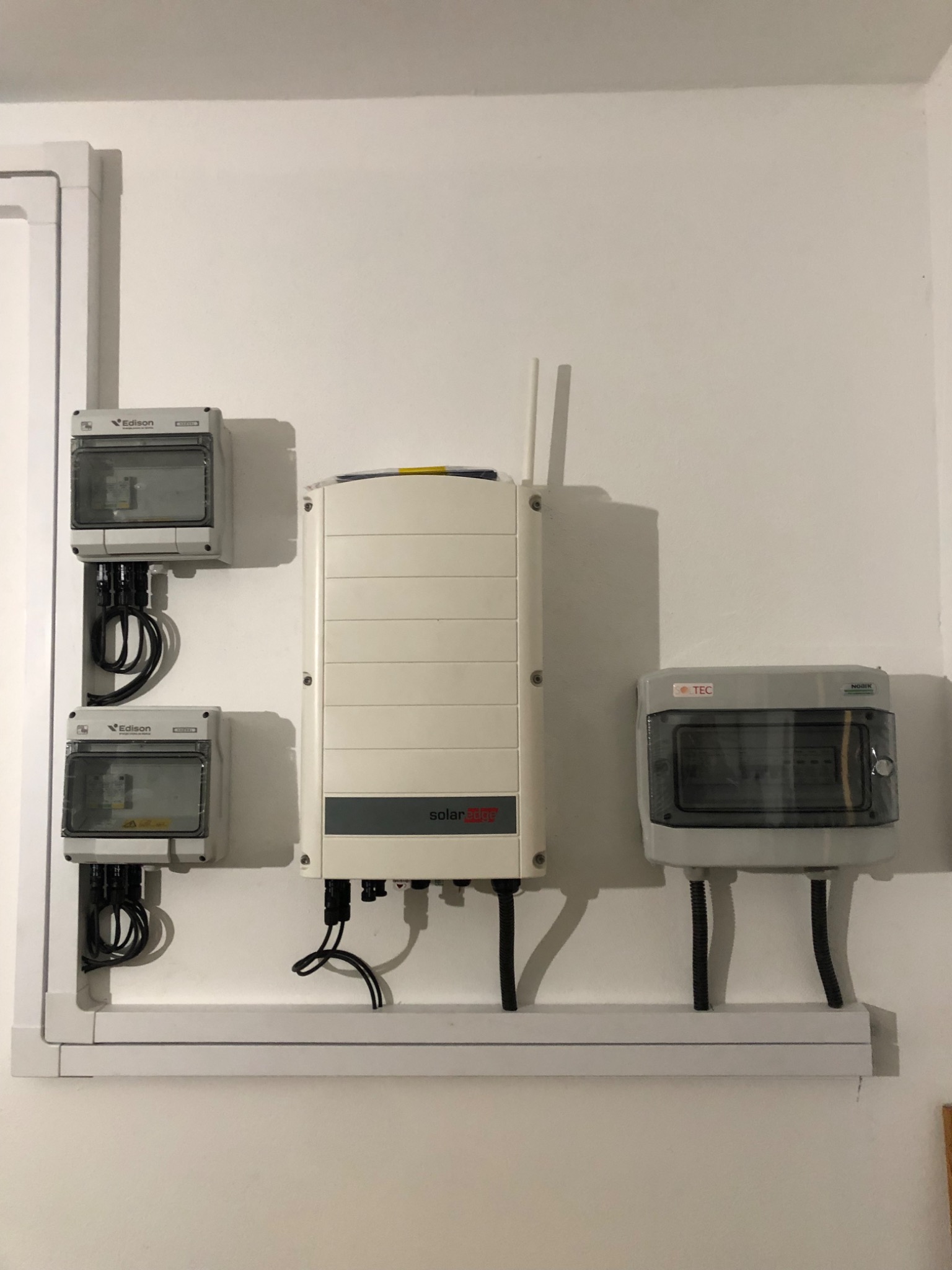 Zamontowany inwerter SolarEdge z dwoma antenami, dwa liczniki energii Edison oraz rozdzielnica TEC, połączone przewodami w listwie przypodłogowej na białej ścianie.