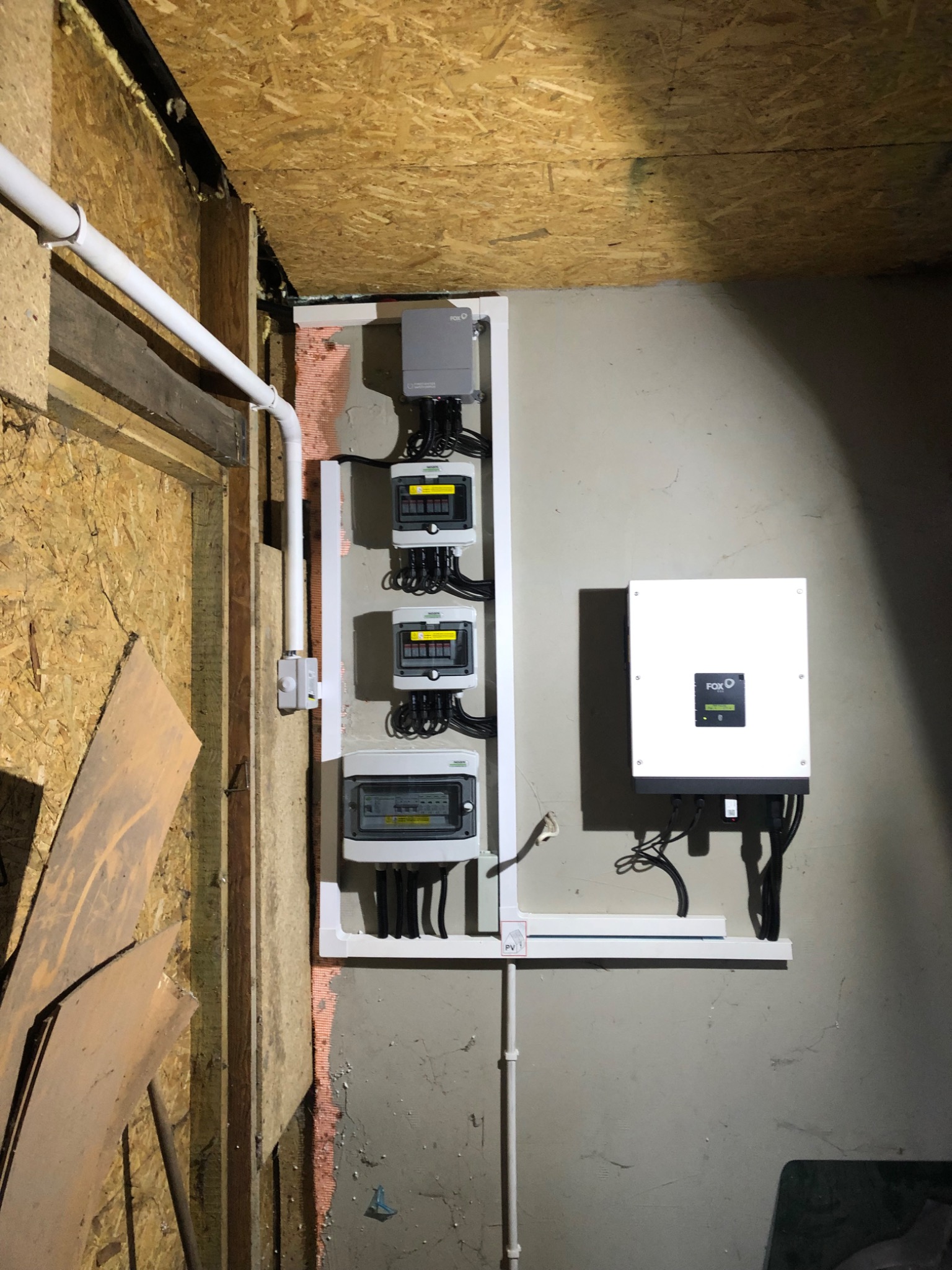 Rozdzielnia elektryczna z trzema licznikami i falownikiem fotowoltaicznym zamontowana na szarej ścianie w surowym pomieszczeniu z boazerią i widoczną instalacją elektryczną.