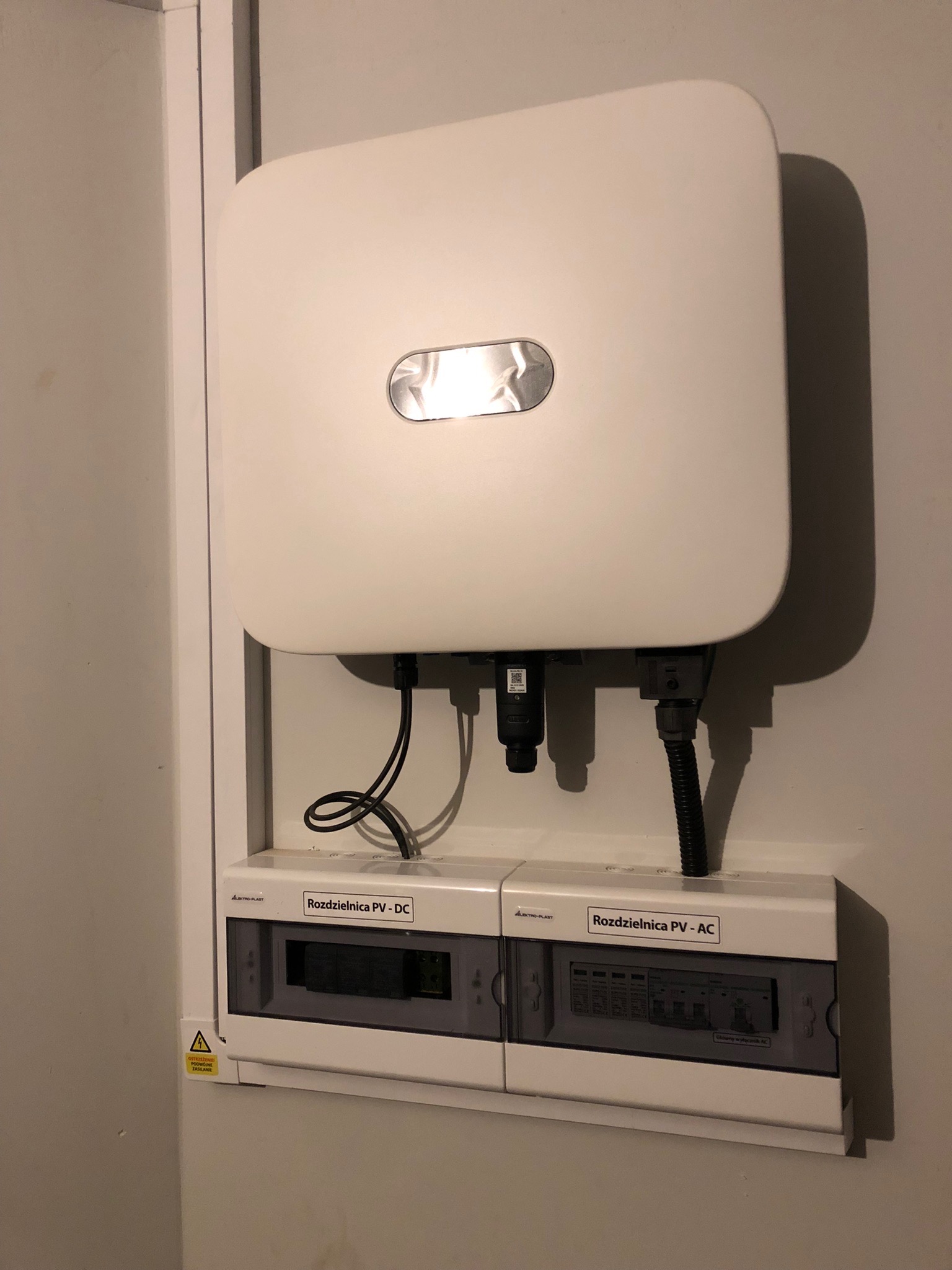 Biały inwerter fotowoltaiczny i dwie rozdzielnice PV-DC oraz PV-AC zamontowane na jasnej ścianie, widoczne czarne kable i żółta naklejka ostrzegawcza.