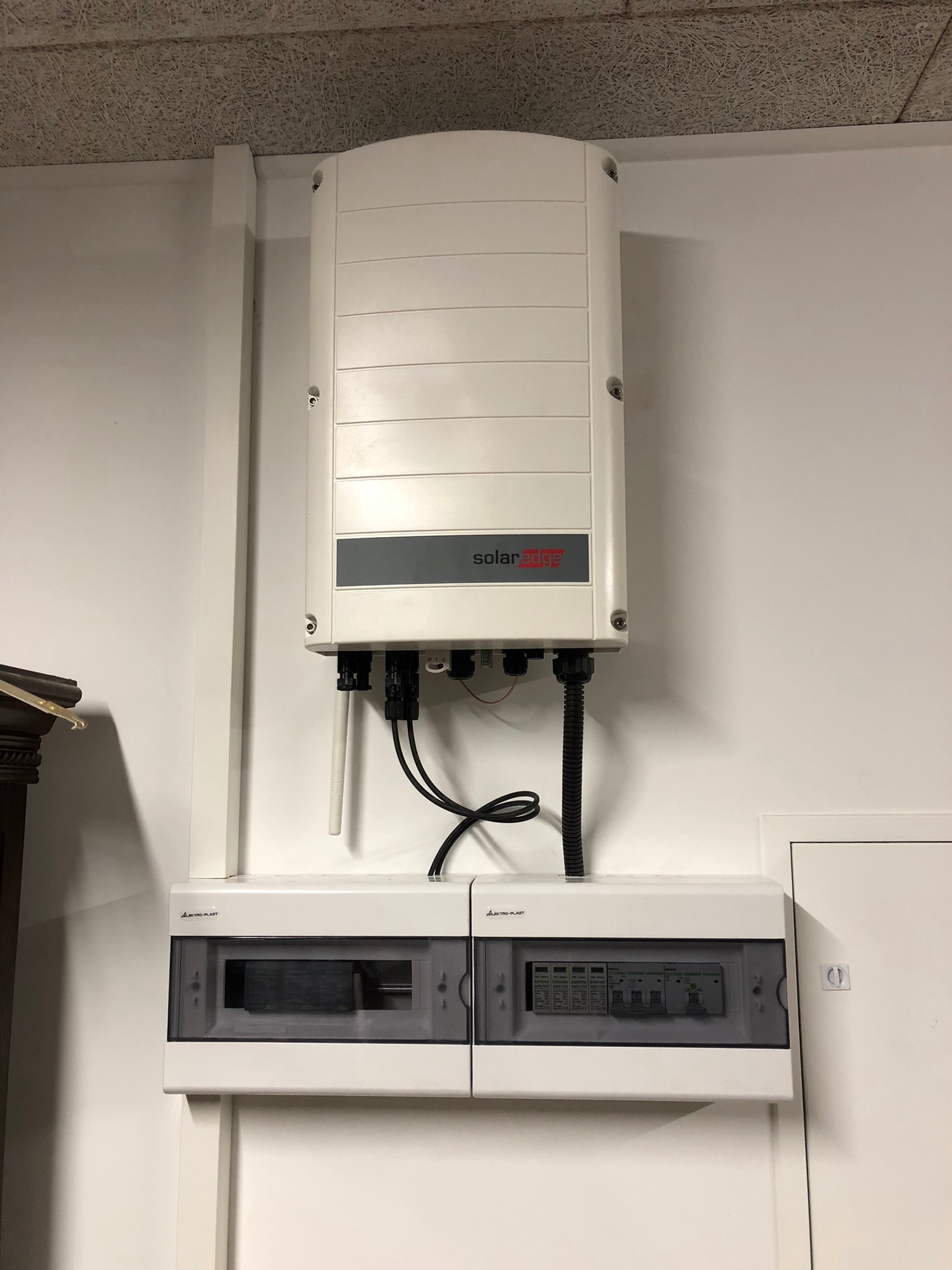 Biały inwerter SolarEdge zamontowany na białej ścianie nad dwiema skrzynkami elektrycznymi z widocznymi bezpiecznikami w szarych obudowach.