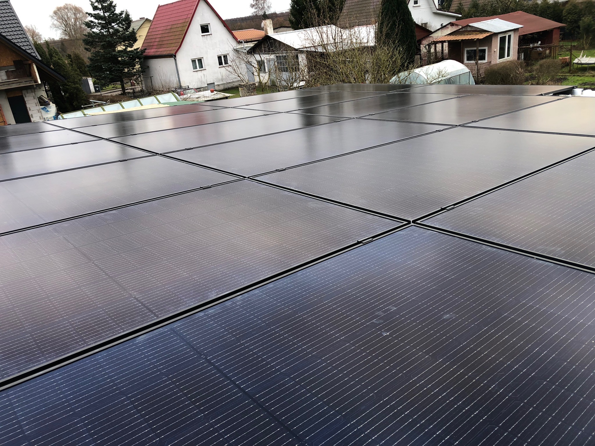 Błyszczące panele słoneczne na dachu domu jednorodzinnego, odbijające pochmurne niebo, z widocznymi sąsiednimi budynkami w tle.