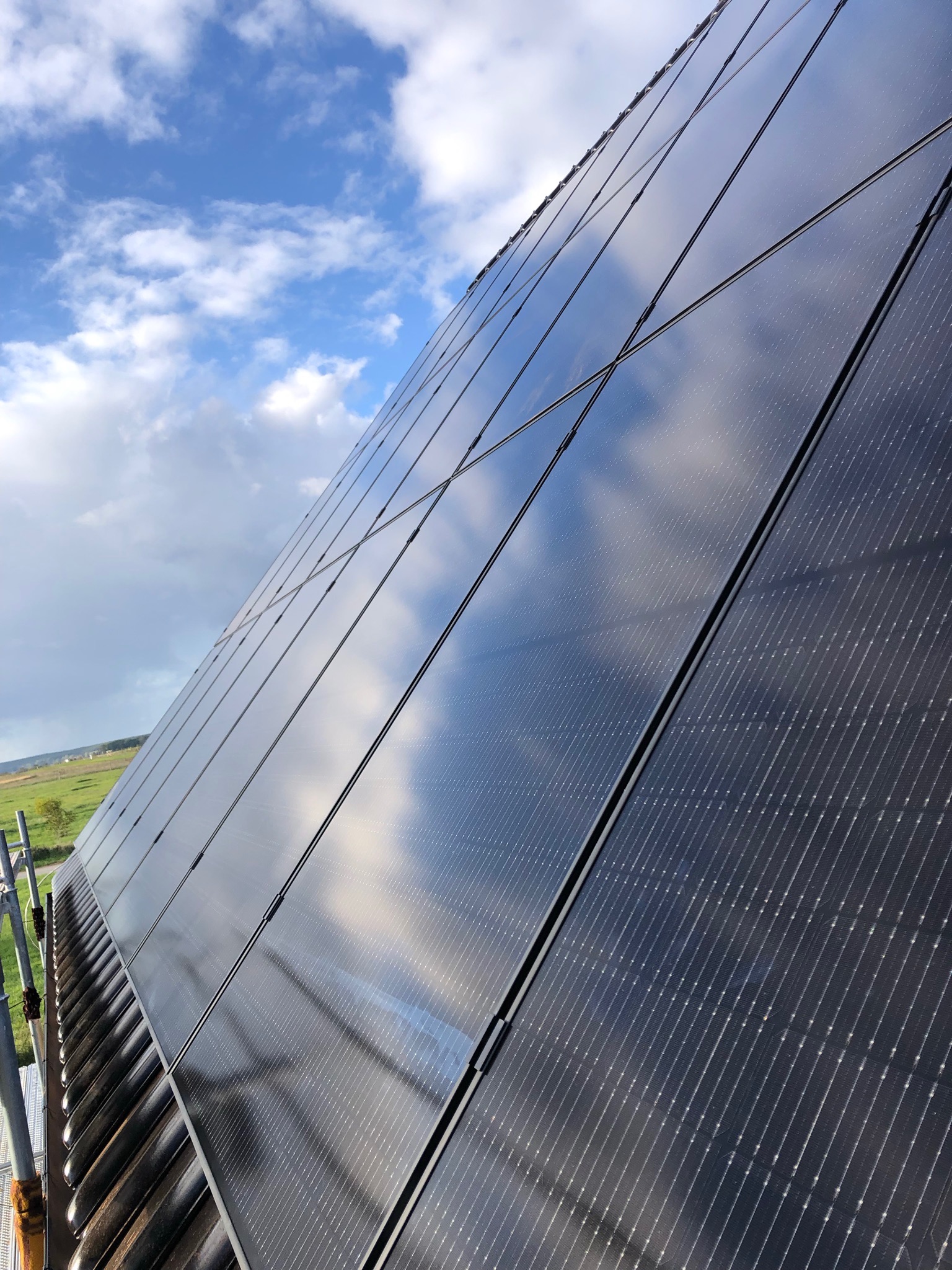 Panele fotowoltaiczne na dachu domu, odbijające błękitne niebo z chmurami, widoczne rusztowanie z boku.