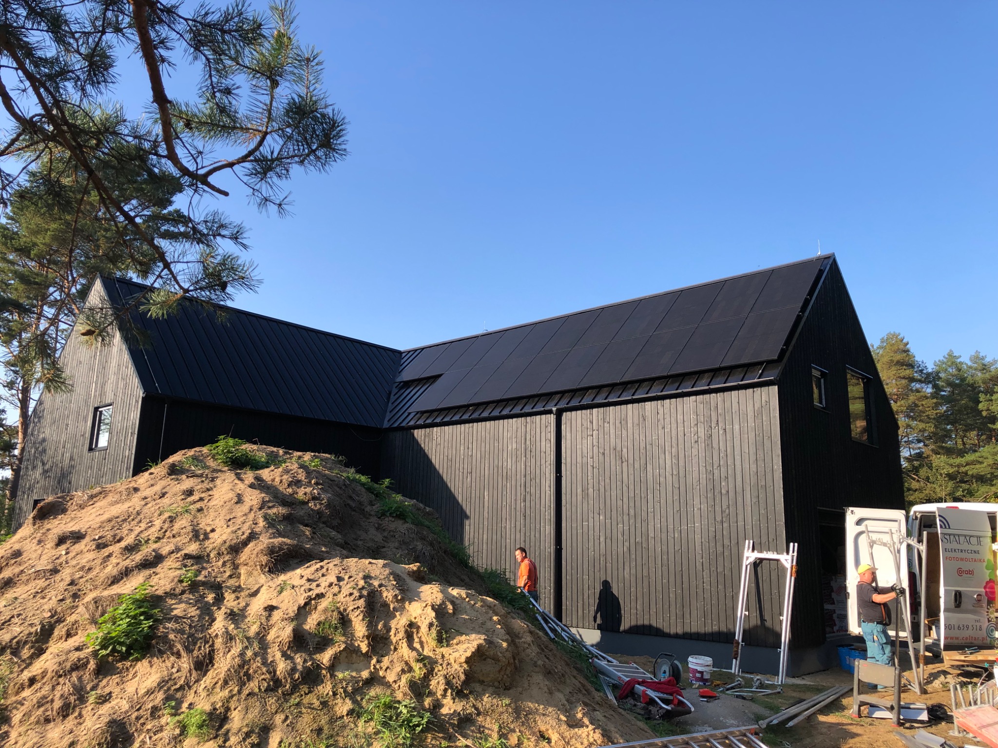Nowoczesny czarny dom z panelami fotowoltaicznymi na dachu, w trakcie montażu, widoczny pracownik i samochód firmy instalacyjnej na tle lasu i błękitnego nieba.