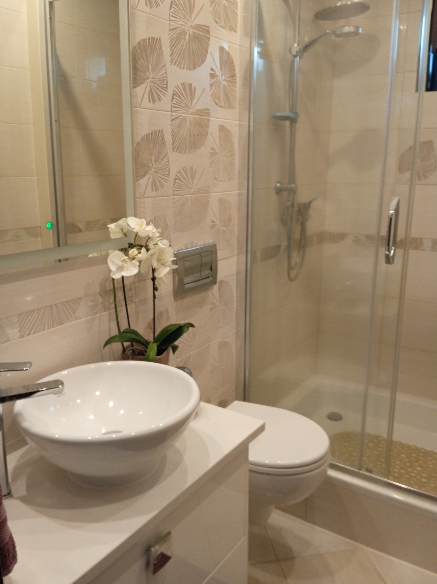 Jasna łazienka z kabiną prysznicową, umywalką nablatową z baterią, toaletą i wzorzystymi płytkami w odcieniach beżu oraz białymi storczykami na blacie.