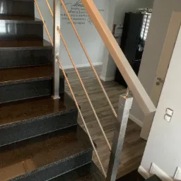 Widok z góry na czarne, polerowane schody z metalową balustradą o kwadratowym przekroju, której pionowe elementy są cieńsze i okrągłe; w tle widoczna ściana z napisem.