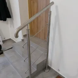 Szczegółowe ujęcie wewnętrznej klatki schodowej z zamontowaną metalową poręczą o minimalistycznym designie, z widocznymi mocowaniami i elementami wykończeniowymi, na tle jasnych ścian i drewnianych...