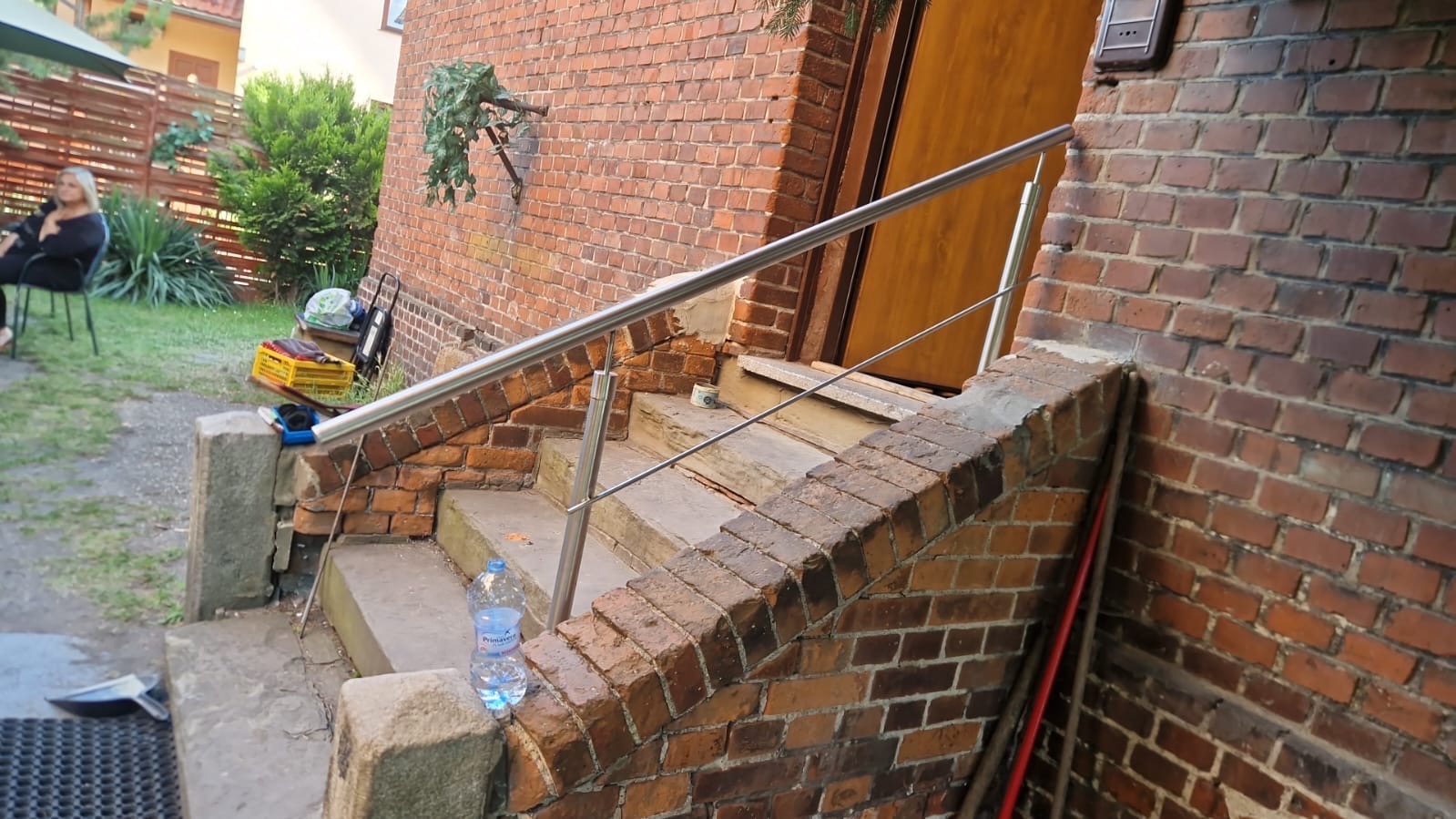 Zewnętrzne schody z cegły z nową, błyszczącą, metalową poręczą, butelka wody na schodach, widok na ogród z siedzącą osobą.