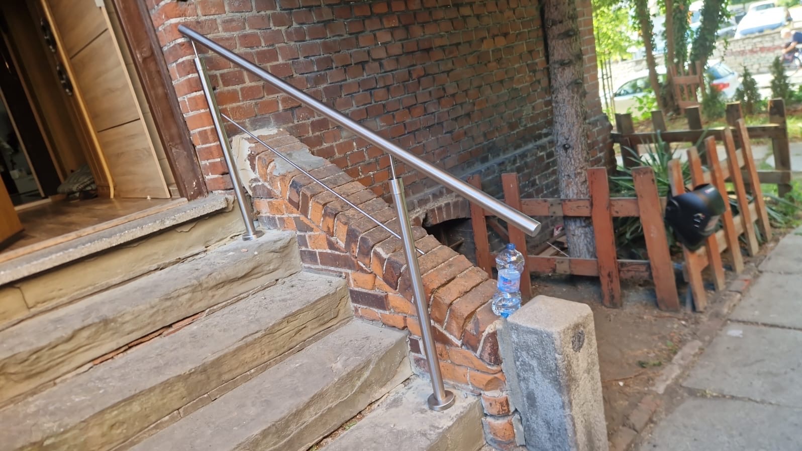 Zewnętrzne schody z kamienia z zamontowaną prostą, metalową poręczą ze stali nierdzewnej. W tle ceglany budynek i drewniane ogrodzenie.