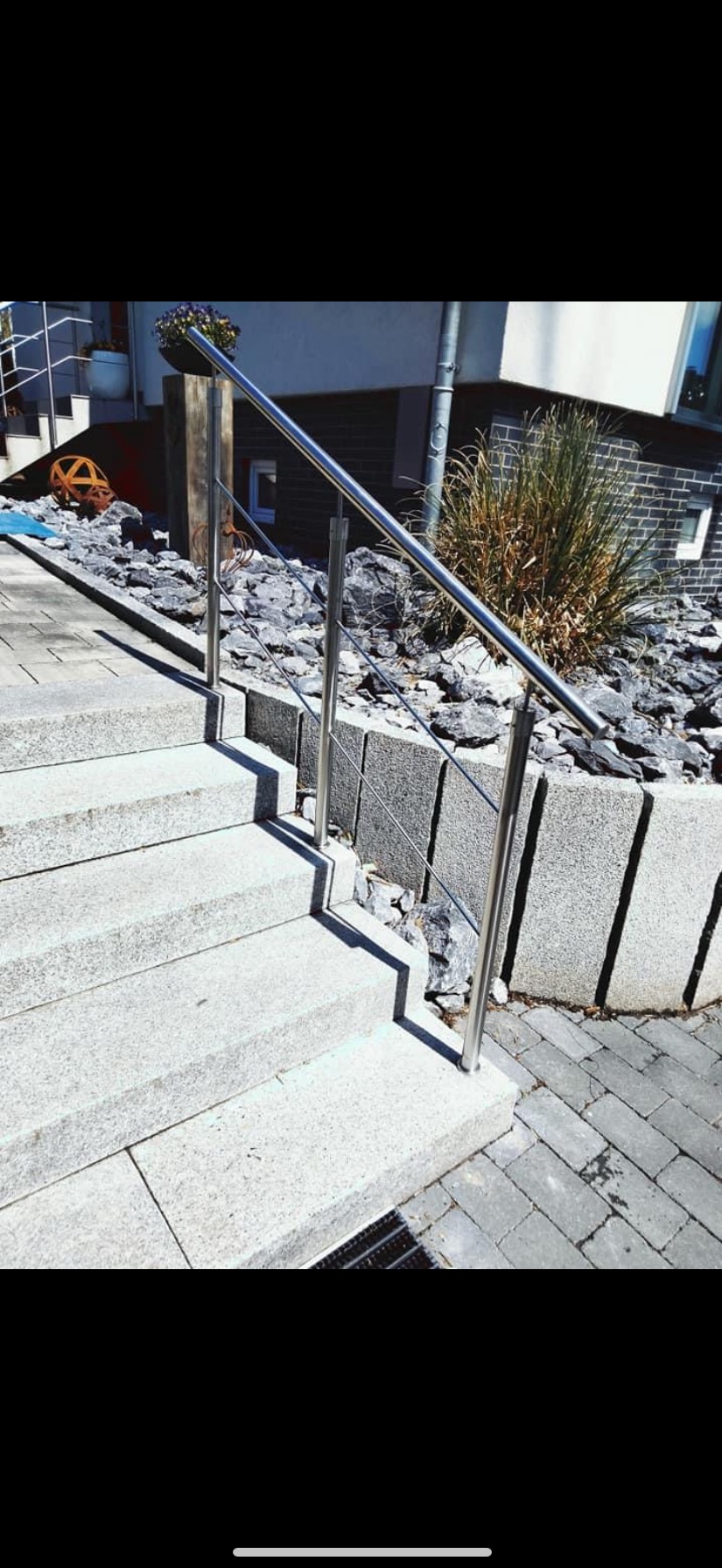 Zewnętrzne schody z jasnego granitu z prostą, nowoczesną balustradą ze stali nierdzewnej i linkami, otoczone kamieniami i niskim murkiem z szarego granitu, z widocznym fragmentem elewacji budynku...