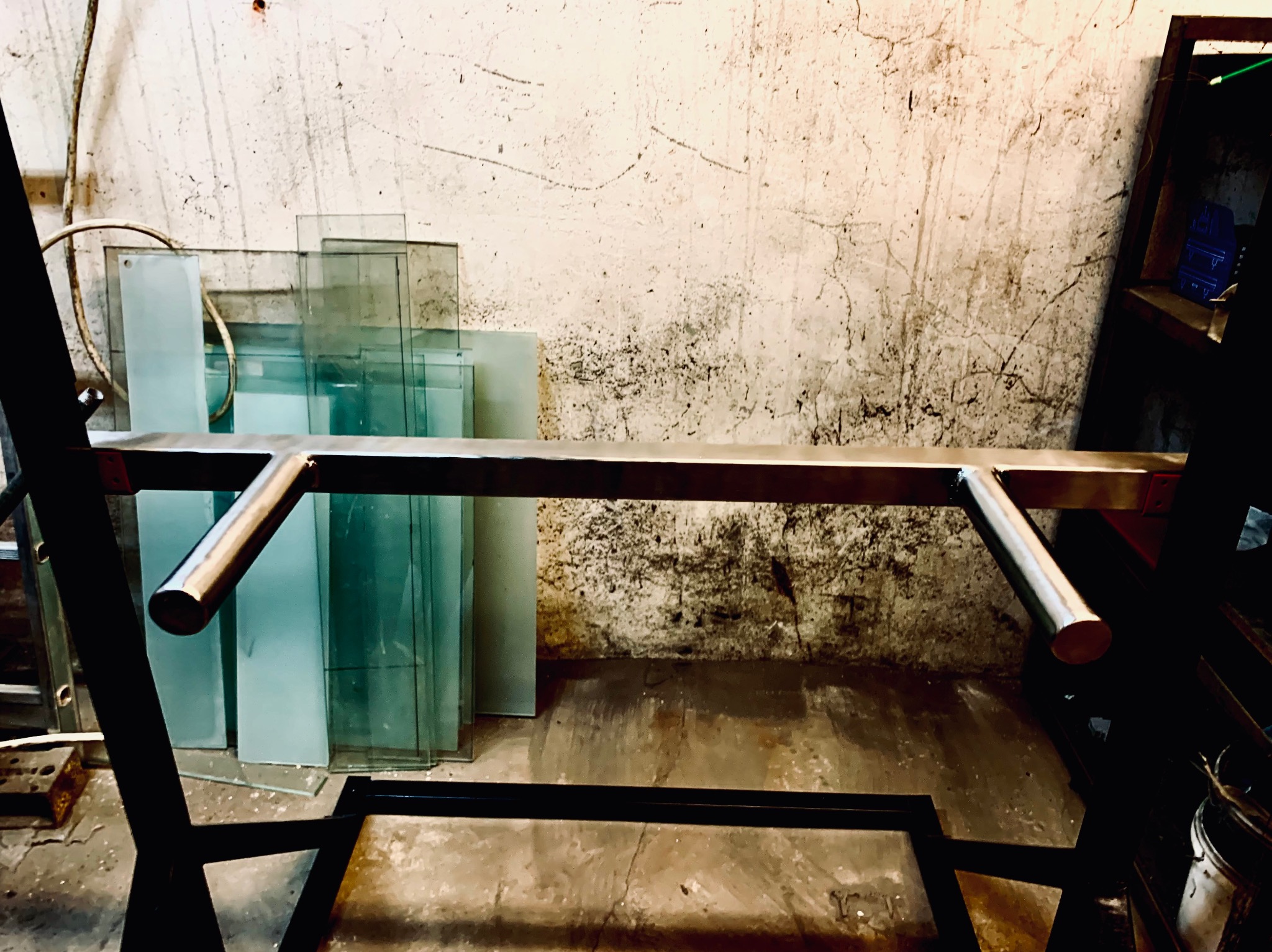 Metalowa konstrukcja w warsztacie, prawdopodobnie element balustrady, z przygotowanymi taflami szkła w tle, widoczna surowa ściana.