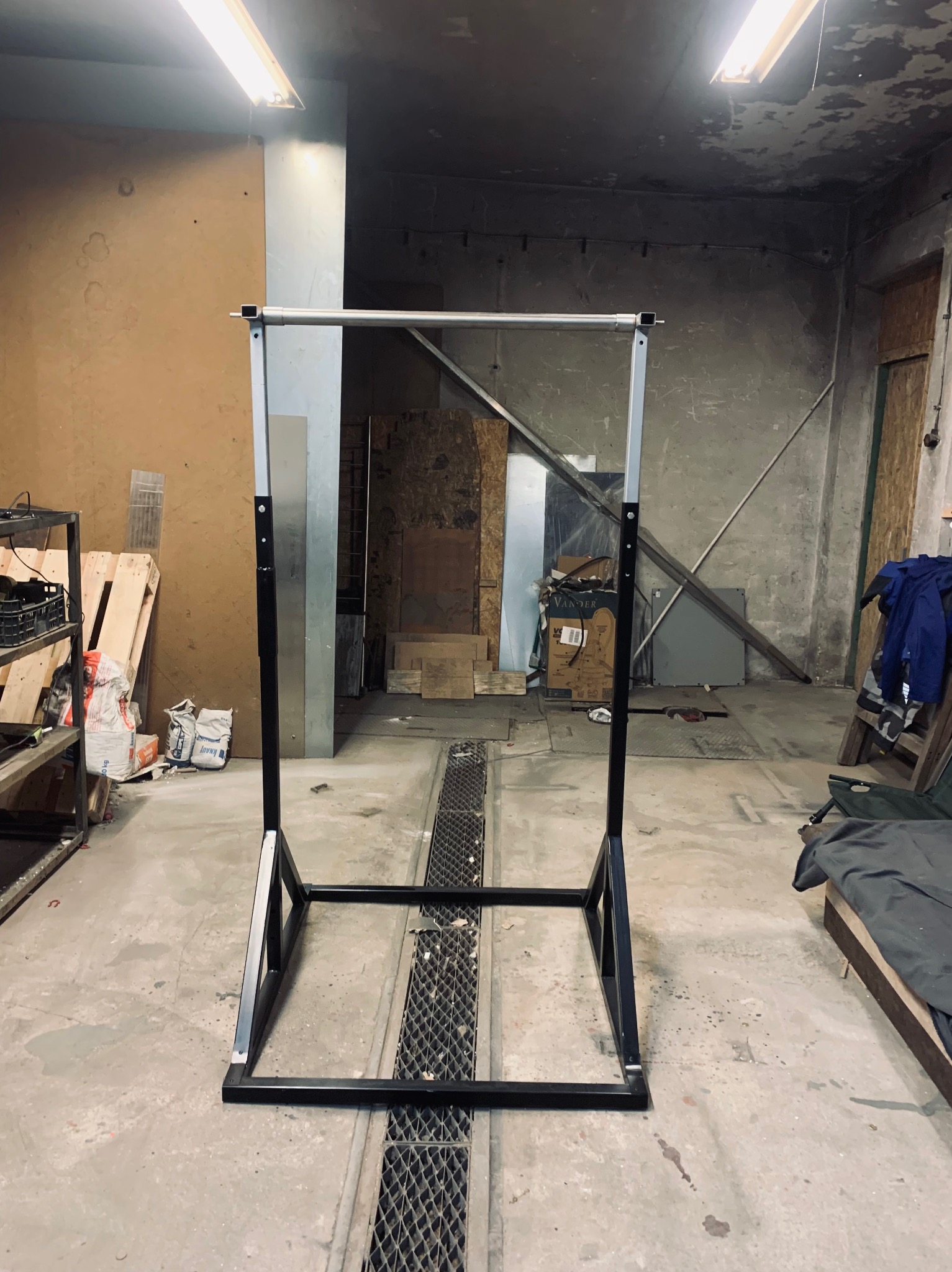 Metalowy stelaż konstrukcyjny z rur, prawdopodobnie do balustrady lub poręczy, stojący w warsztacie z widocznymi materiałami budowlanymi i narzędziami.