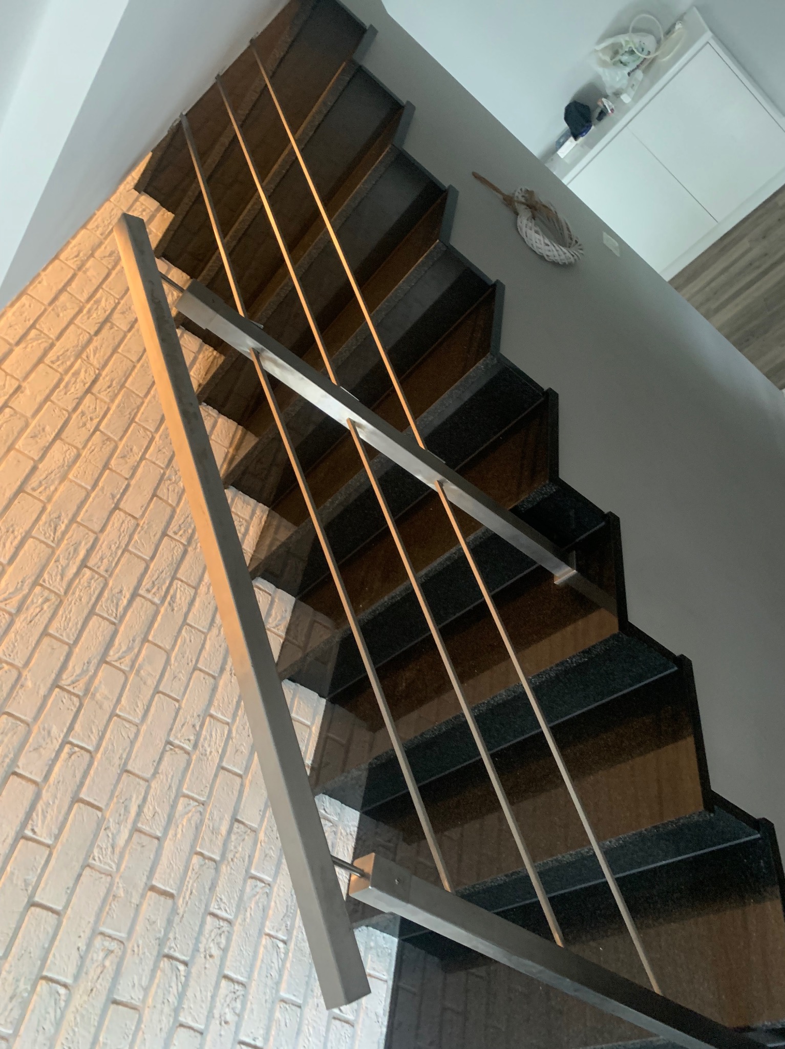 Metalowe schody z czarnymi stopniami i minimalistyczną szarą balustradą z poziomych prętów, widok z dołu, ściana z białej cegły dekoracyjnej po lewej stronie.