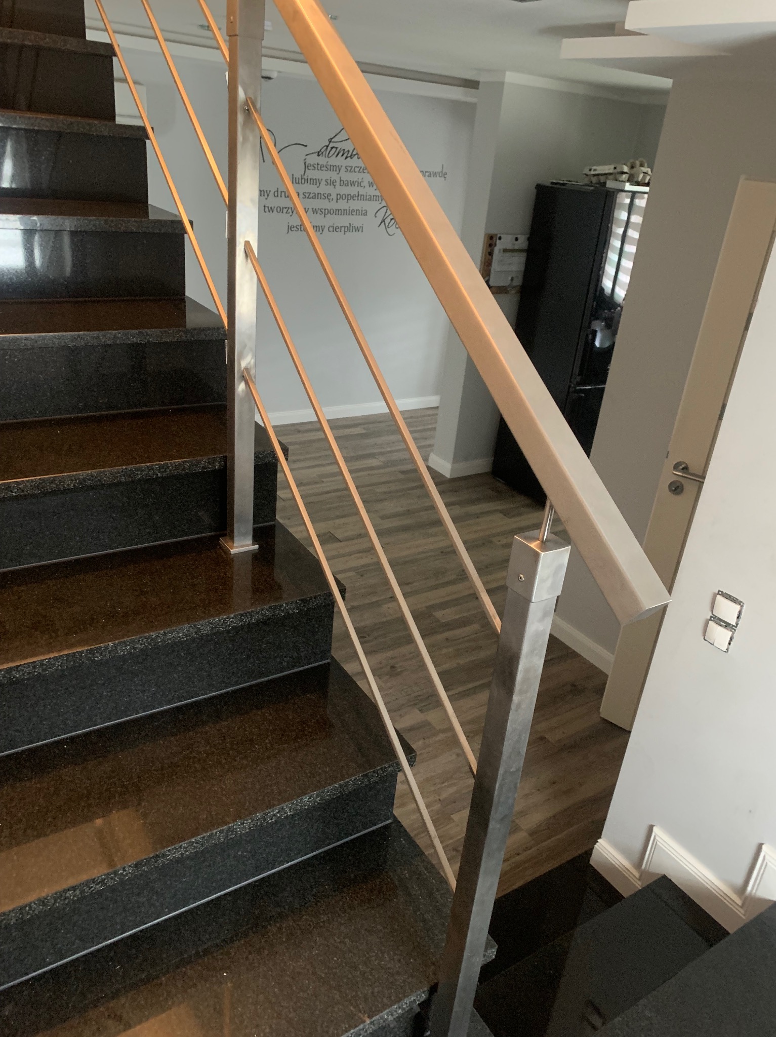 Widok z góry na czarne, polerowane schody z metalową balustradą o kwadratowym przekroju, której pionowe elementy są cieńsze i okrągłe; w tle widoczna ściana z napisem.