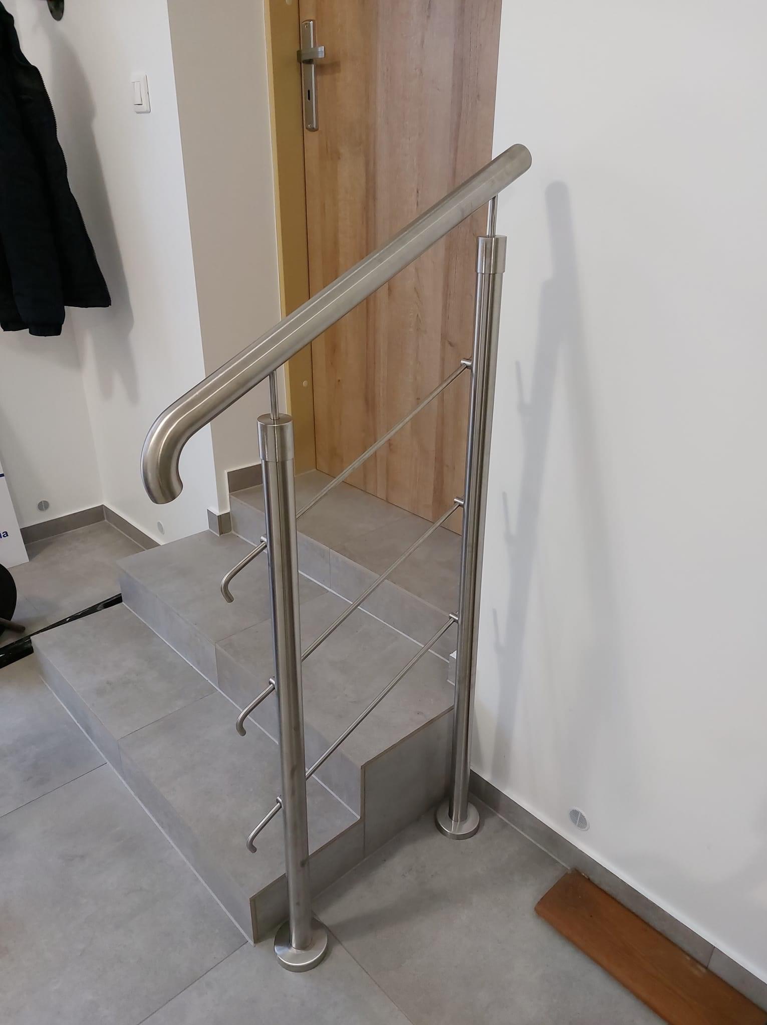 Szczegółowe ujęcie wewnętrznej klatki schodowej z zamontowaną metalową poręczą o minimalistycznym designie, z widocznymi mocowaniami i elementami wykończeniowymi, na tle jasnych ścian i drewnianych...