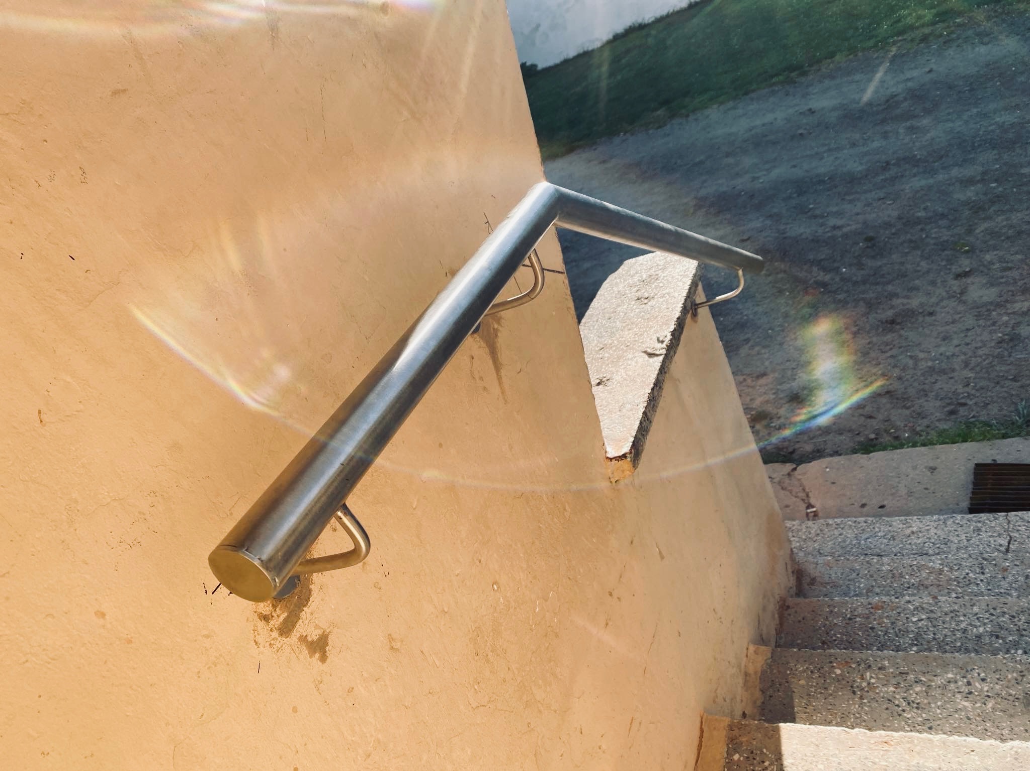 Metalowa poręcz zamontowana na ścianie obok betonowych schodów zewnętrznych, z efektem tęczy spowodowanym przez odbicie światła.