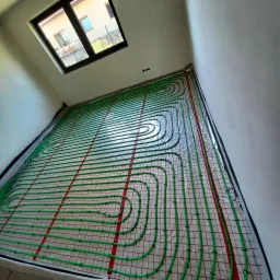 Zielone rury ogrzewania podłogowego ułożone na folii z siatką, widok z góry na instalację przed zalaniem posadzki, okno dachowe w tle.