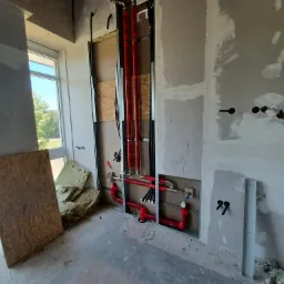 Wnętrze w trakcie remontu z widocznymi czerwonymi rurami instalacji grzewczej w ścianie, izolacją i płytą OSB przy oknie.