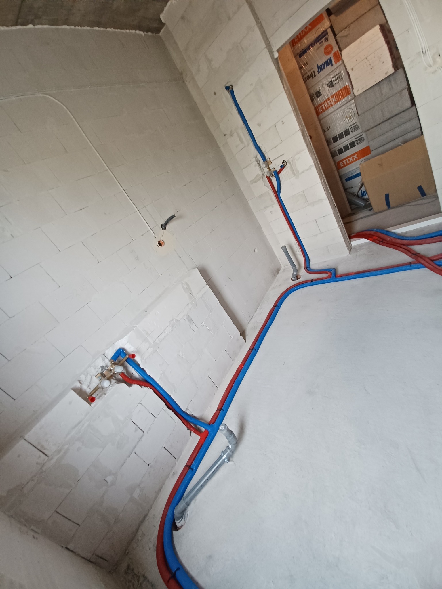Surowe wnętrze w trakcie prac hydraulicznych: czerwono-niebieskie rury prowadzone po ścianach z betonu komórkowego i wylewce, widoczne elementy instalacji i skrzynka z materiałami budowlanymi w tle.
