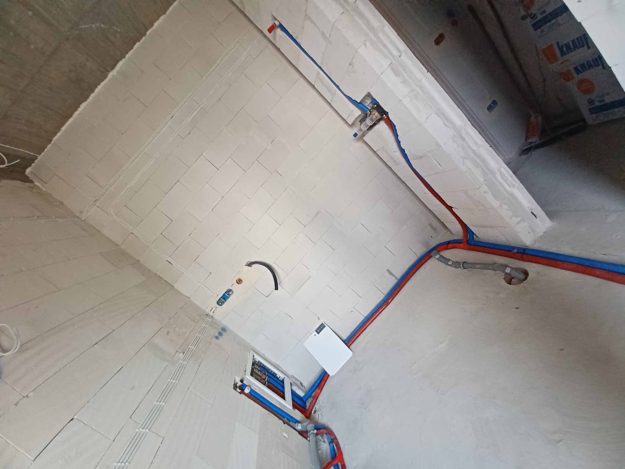 Wnętrze w trakcie instalacji hydraulicznej i grzewczej: widoczne rury PEX w kolorach czerwonym i niebieskim prowadzone po ścianach z bloczków, podłączenia do rozdzielaczy i punktów poboru wody...