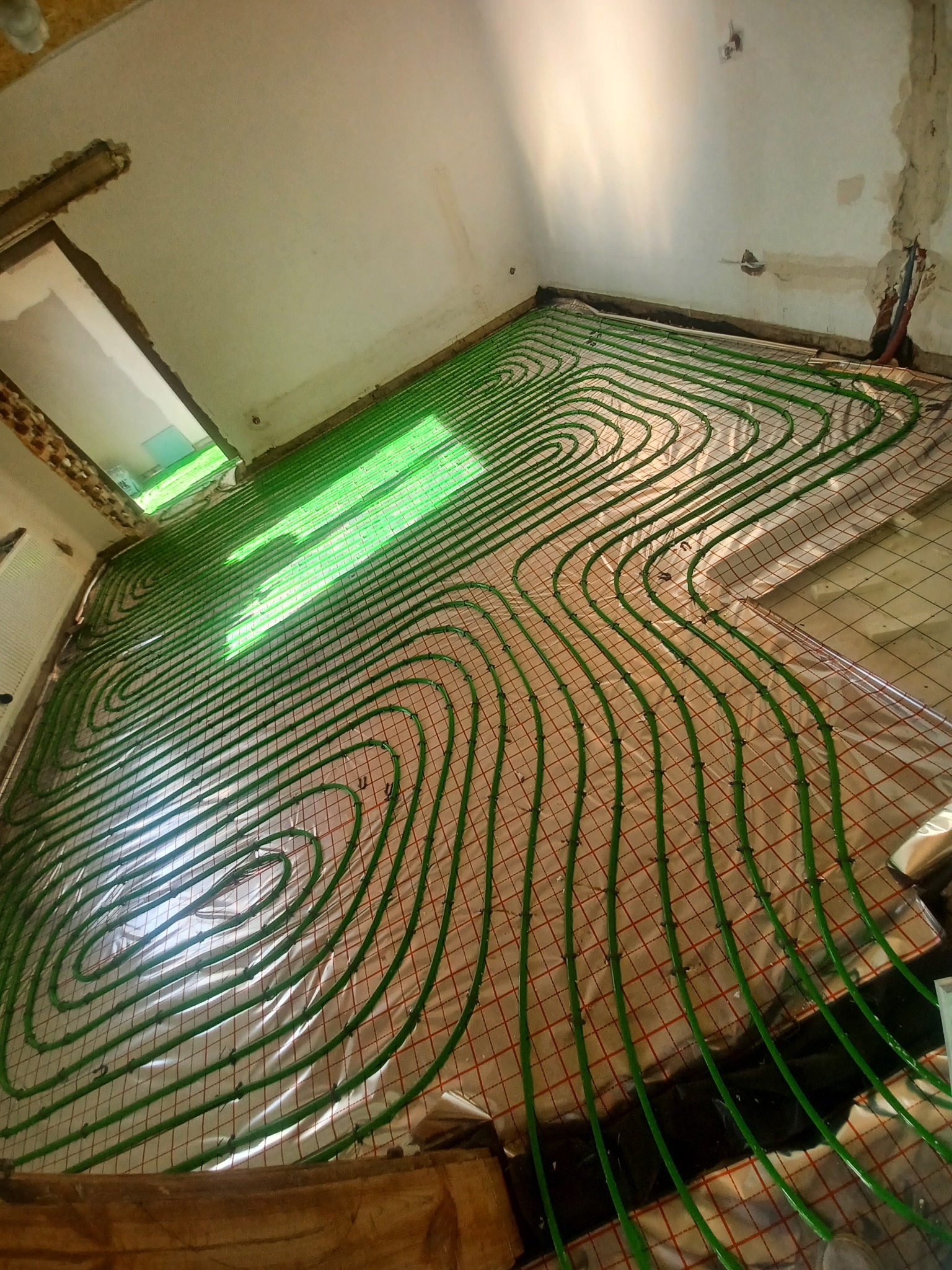 Zielone rurki ogrzewania podłogowego ułożone spiralnie na siatce w surowym pomieszczeniu, widoczne fragmenty ściany i otworu okiennego w trakcie remontu.