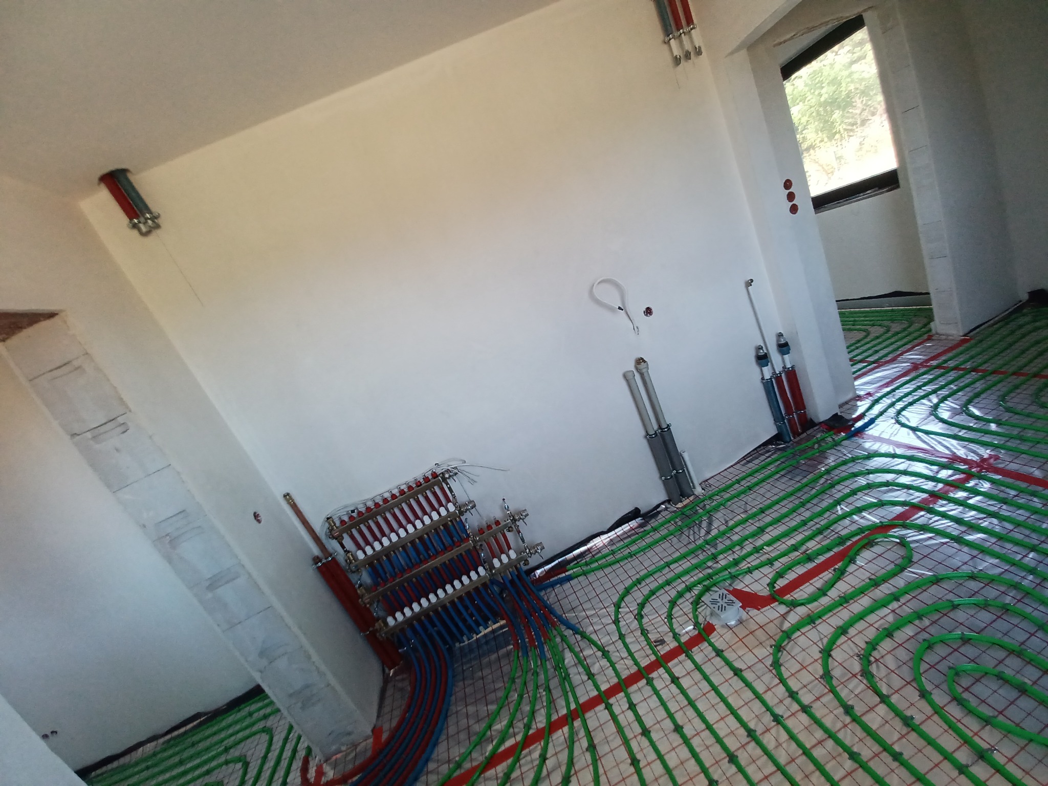 Rozdzielacz ogrzewania podłogowego z rurami ułożonymi na folii izolacyjnej w surowym stanie, widoczne przyłącza do instalacji centralnego ogrzewania.