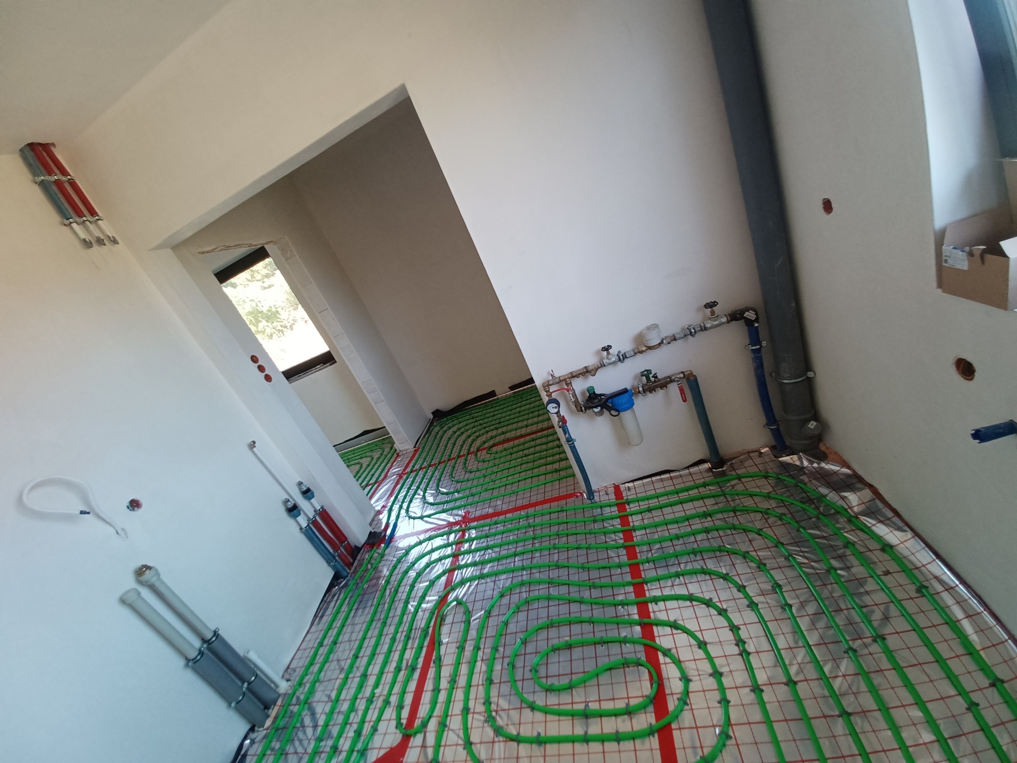 Zielone rurki ogrzewania podłogowego ułożone na folii odblaskowej w nowym, jasnym pomieszczeniu. Widoczne również elementy instalacji wodnej na ścianach.