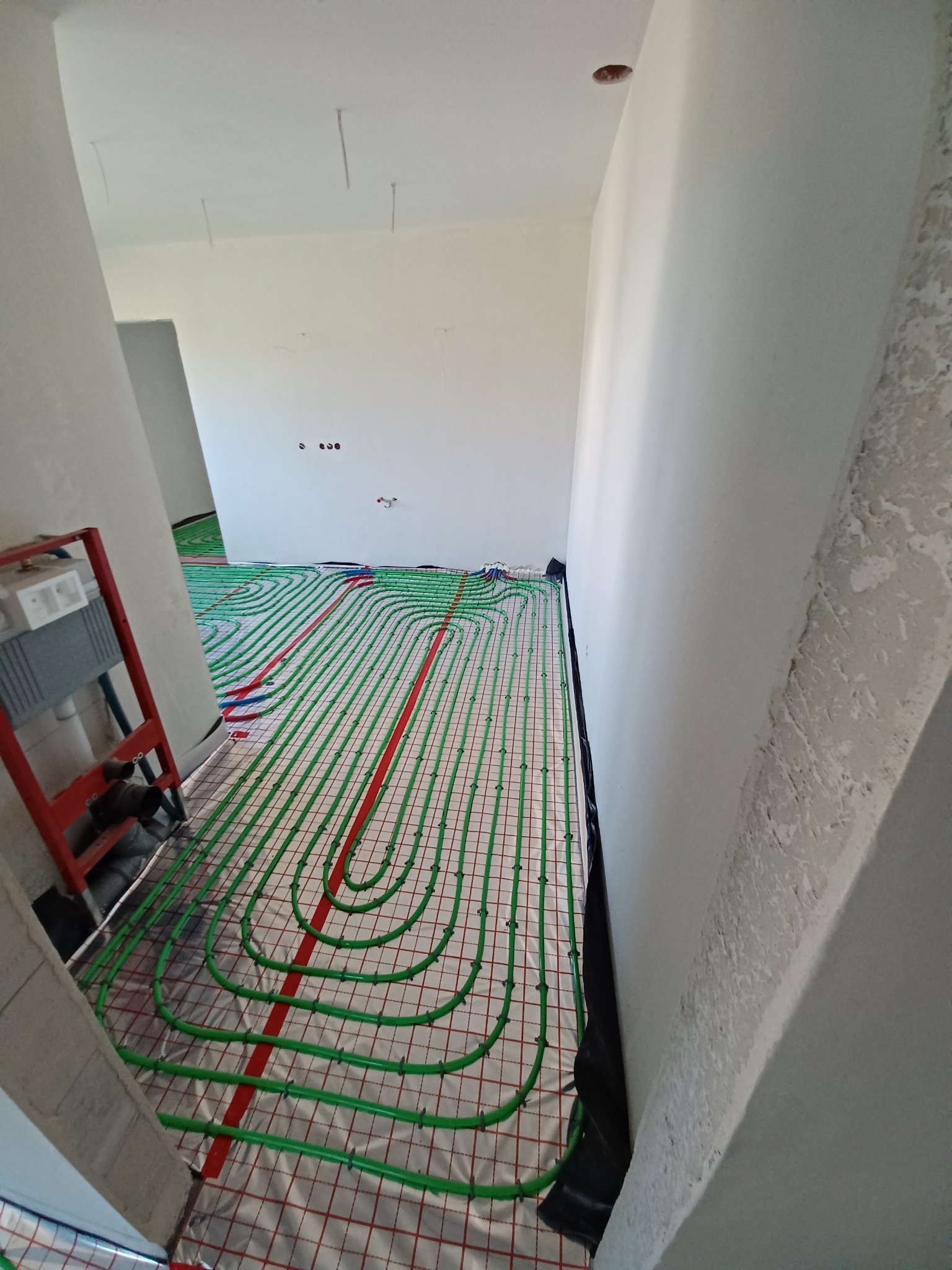 Układanie zielonych rur ogrzewania podłogowego na folii izolacyjnej z czerwoną siatką w surowym wnętrzu z widoczną czerwoną drabinką i instalacją sanitarną przy ścianie.