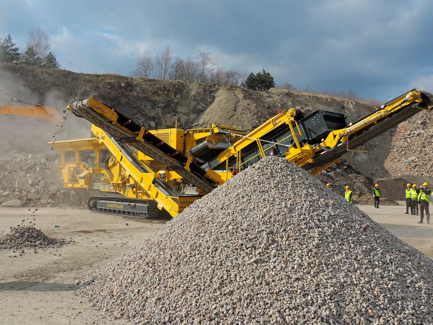 Żółta kruszarka Keestrack na gąsienicach przerabia gruz na żwir, z tyłu hałda gruzu i grupa osób w kamizelkach odblaskowych.