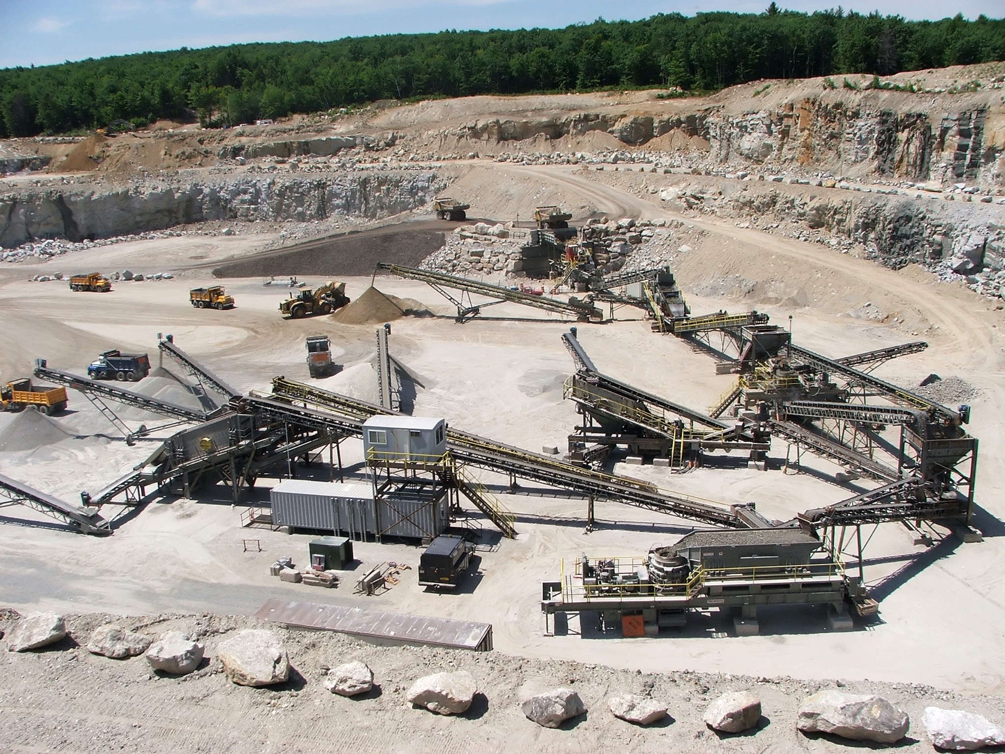 Widok z góry na rozległą kopalnię odkrywkową z ciężkim sprzętem, w tym koparkami, ciężarówkami i systemem przenośników taśmowych, zalesione wzgórza w tle.