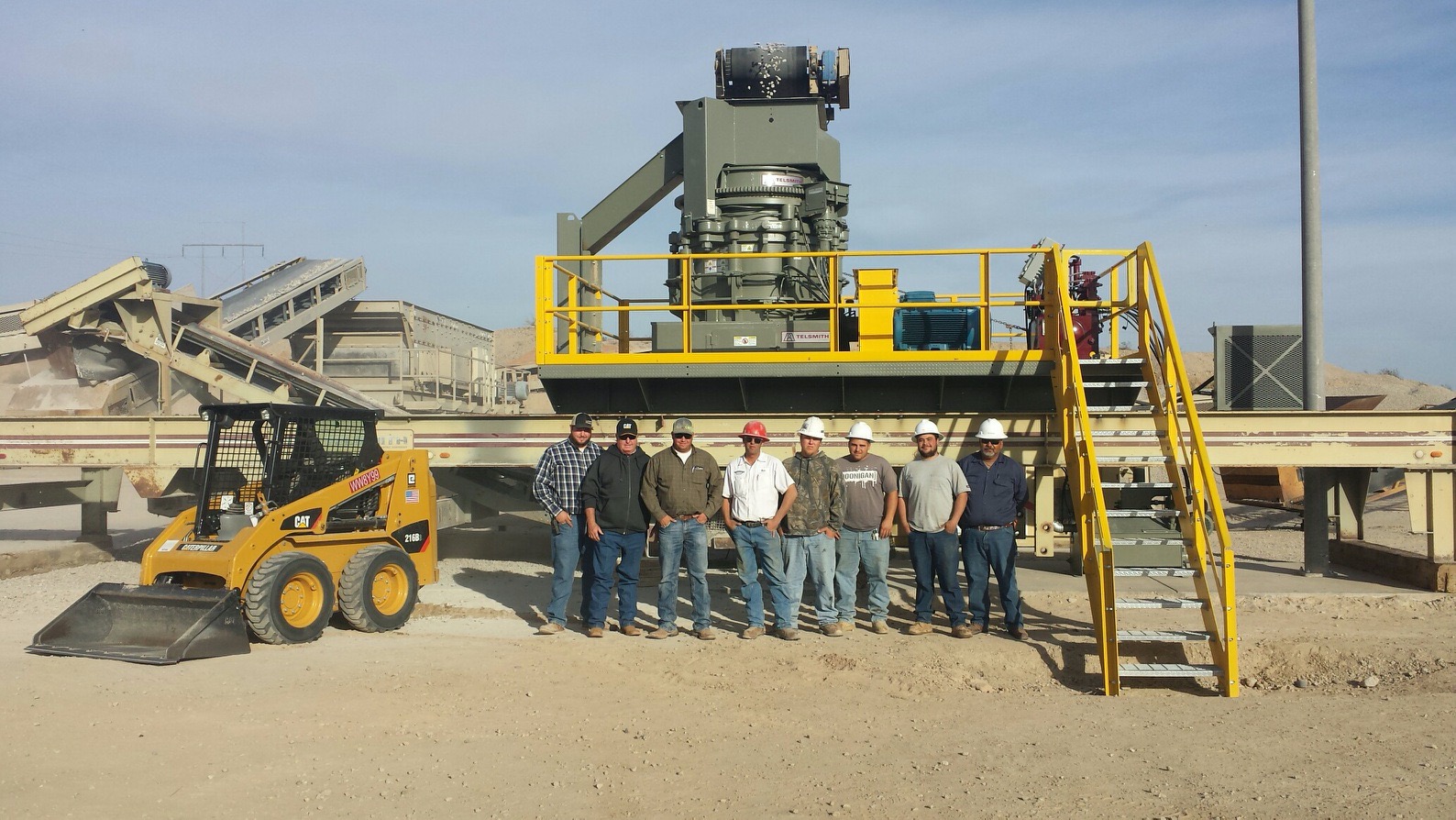 Grupa ośmiu pracowników w kaskach pozuje na tle kruszarki do kamienia marki Telsmith i ładowarki CAT 216B na terenie budowy.