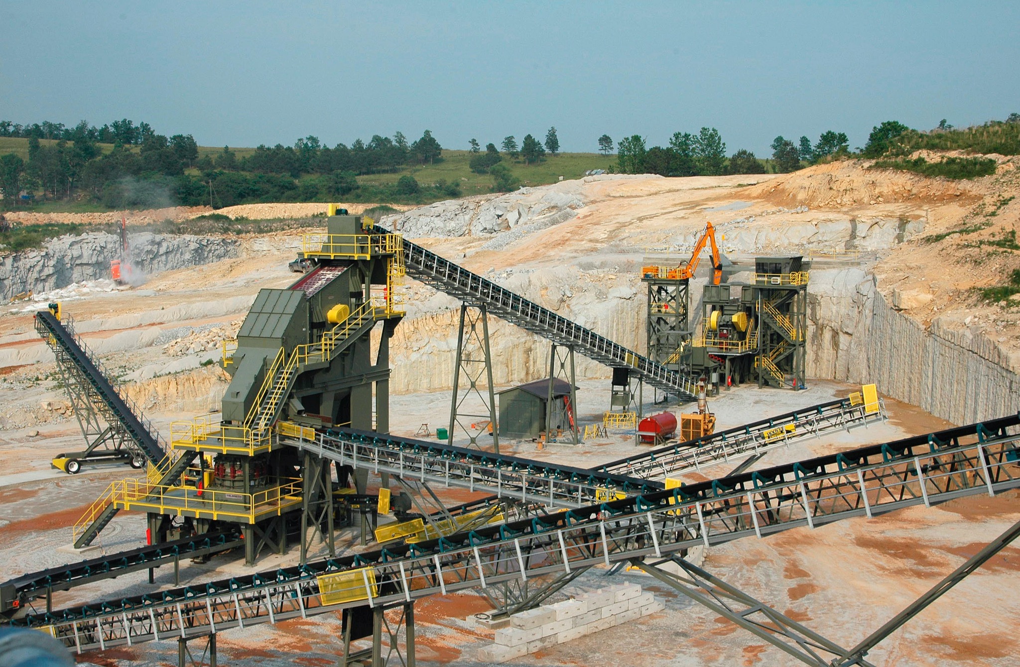 Widok z góry na rozległą kopalnię odkrywkową z systemem przenośników taśmowych i maszynami do kruszenia kamienia na tle zielonych wzgórz.